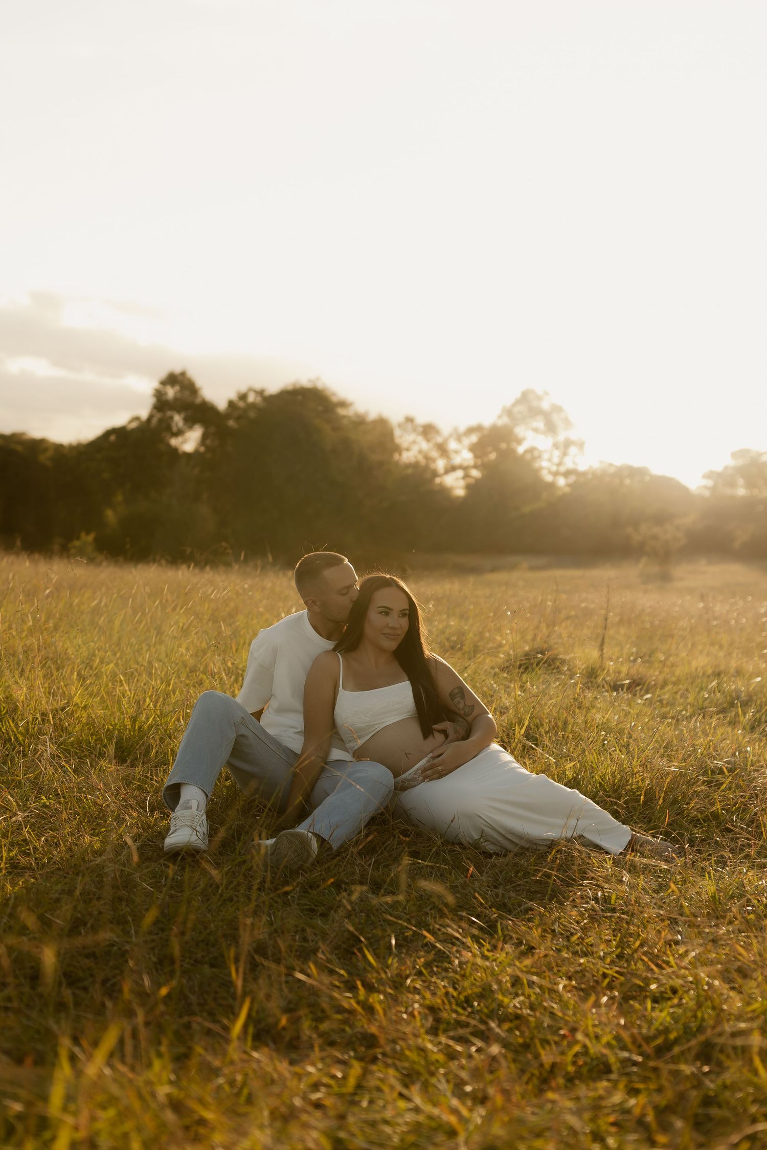 Couple sitting in golden field at sunset, man kissing pregnant woman's cheek while holding her baby bump.