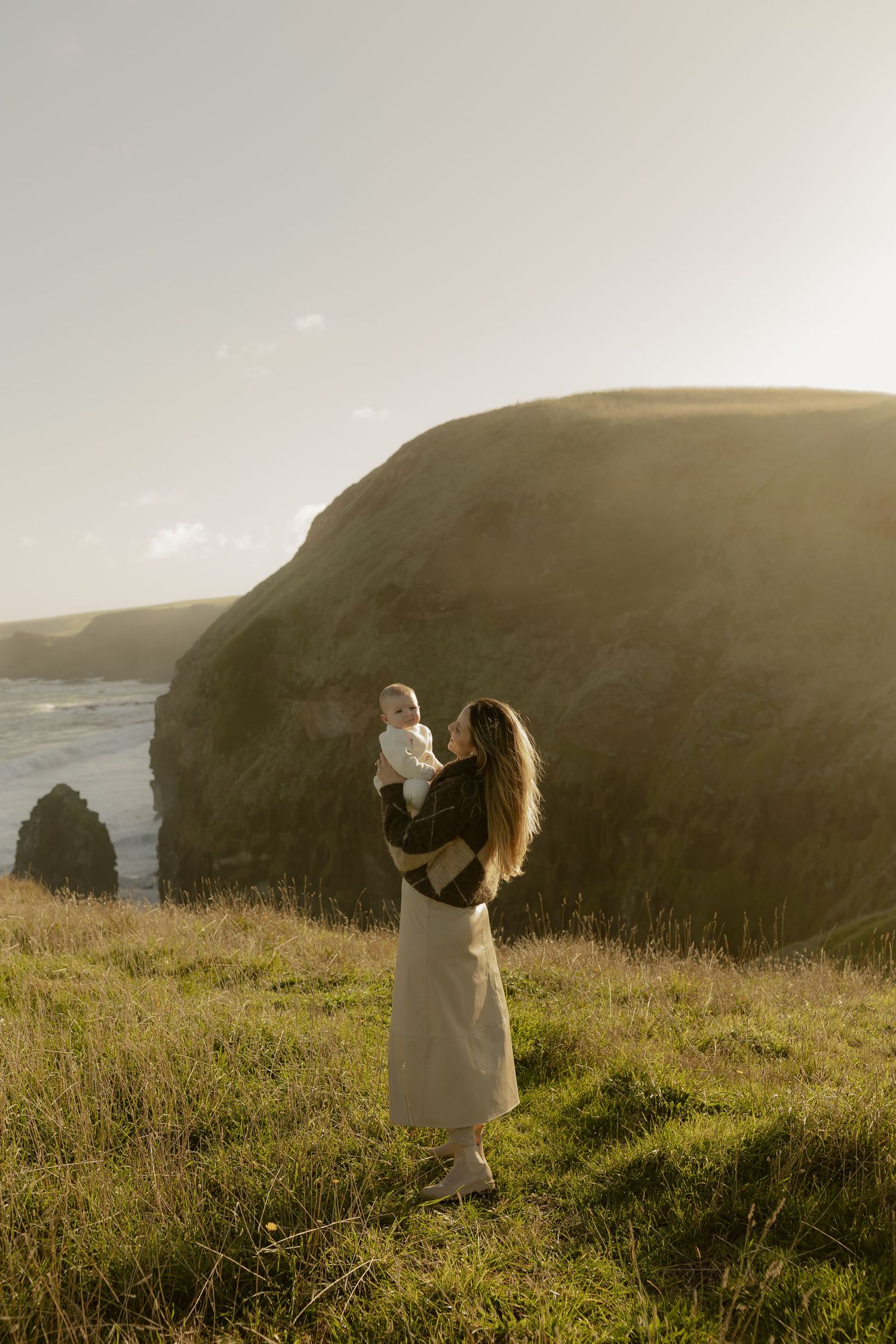 A mother holds her baby on a scenic coastal cliff at golden hour with green hills and ocean views.