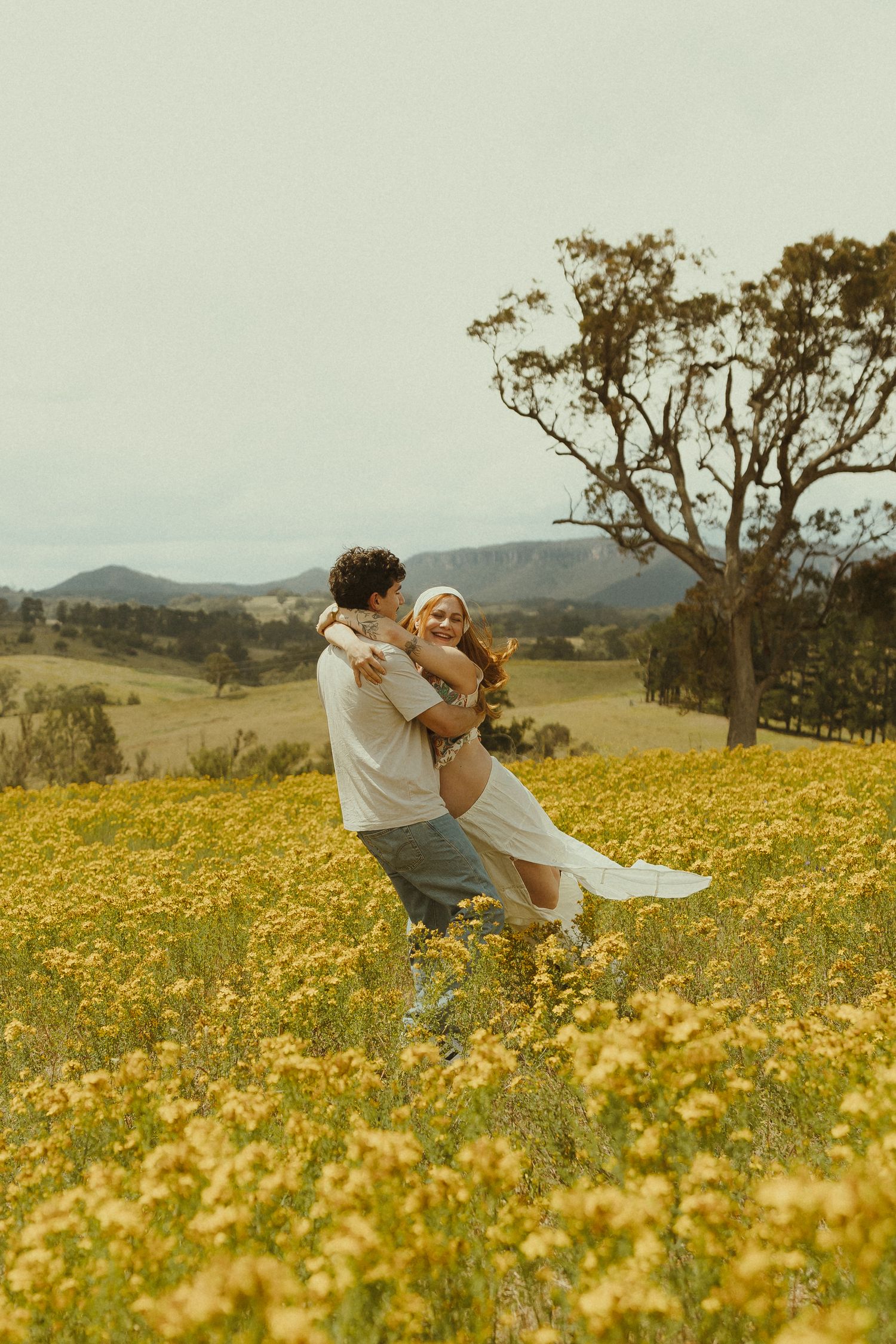A romantic couple embraces in a meadow of yellow wildflowers with mountains in the distance.