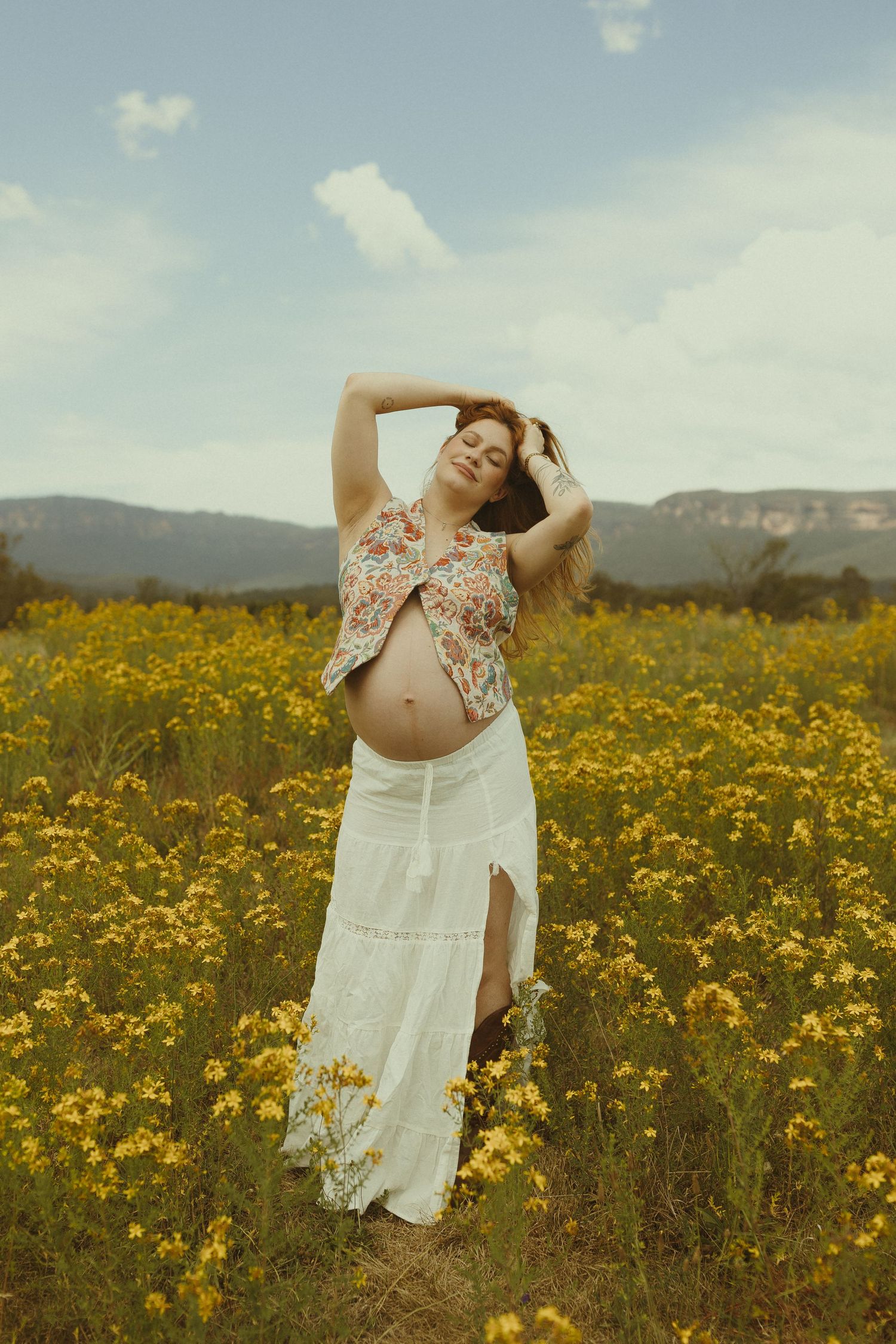 Woman in white dress poses gracefully in a field of yellow wildflowers against mountain backdrop.