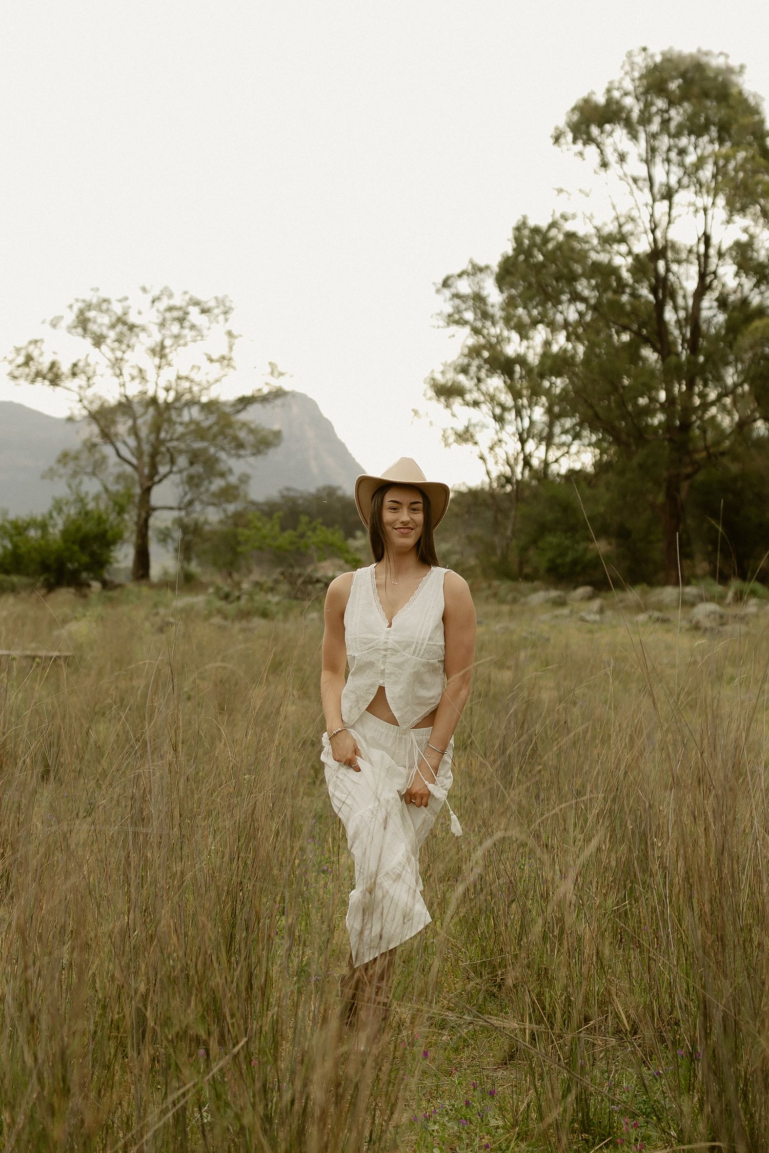 A figure in white twirls playfully through tall grass with mountains in the background.