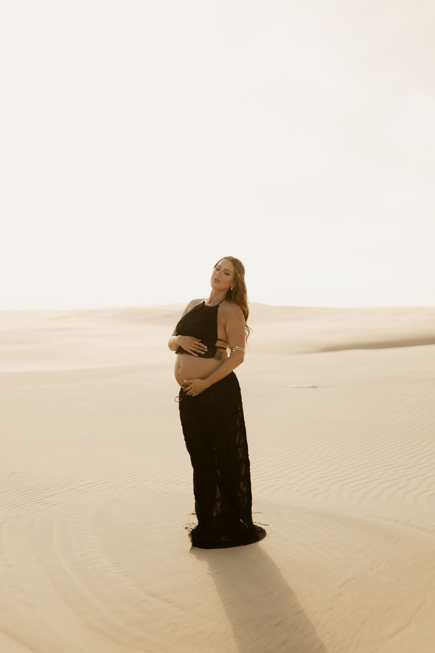 Figure in black dress poses dramatically against sweeping sand dunes in sepia toned desert landscape.