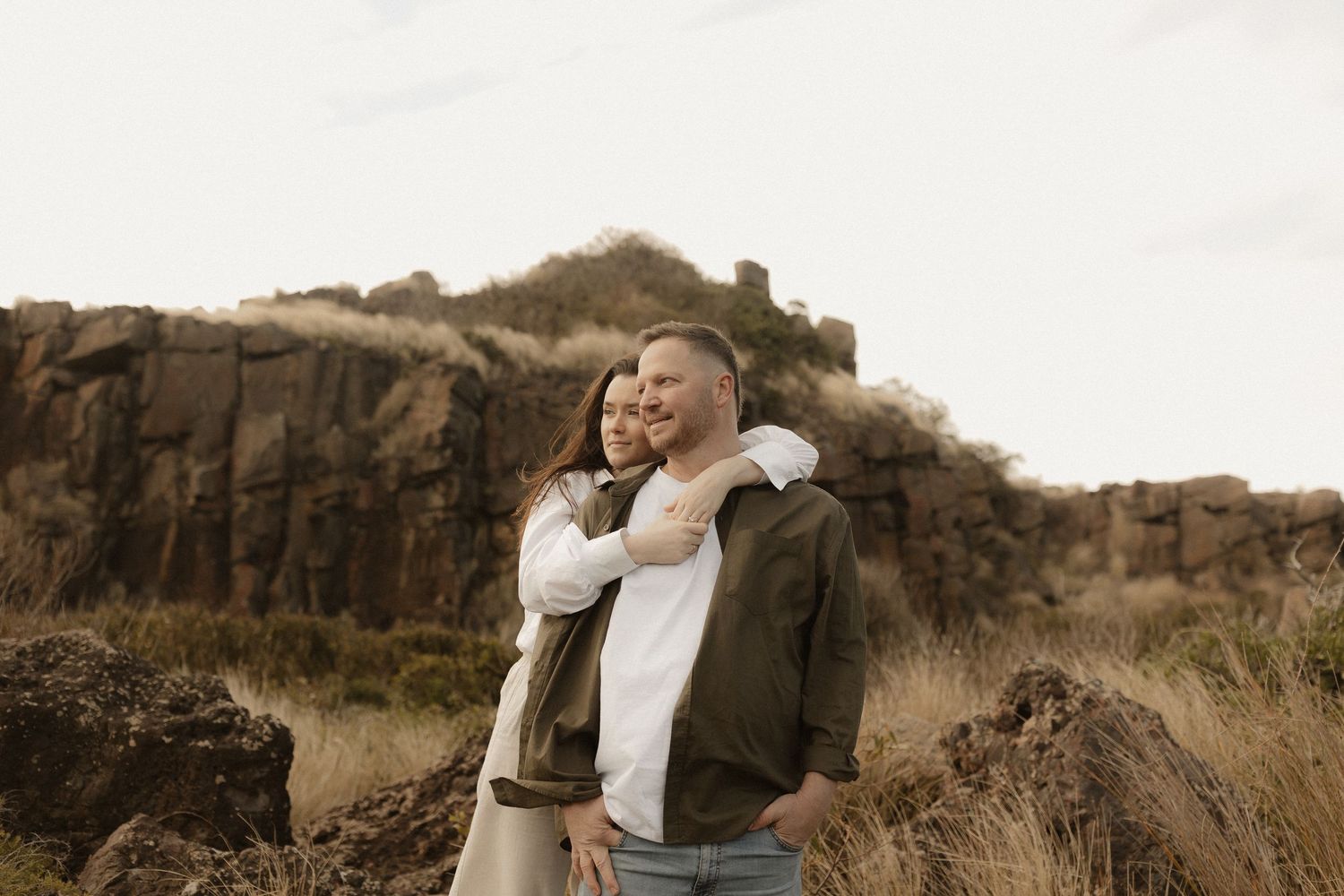Couple shares intimate moment on rocky cliffside overlooking ocean during sunset photoshoot.