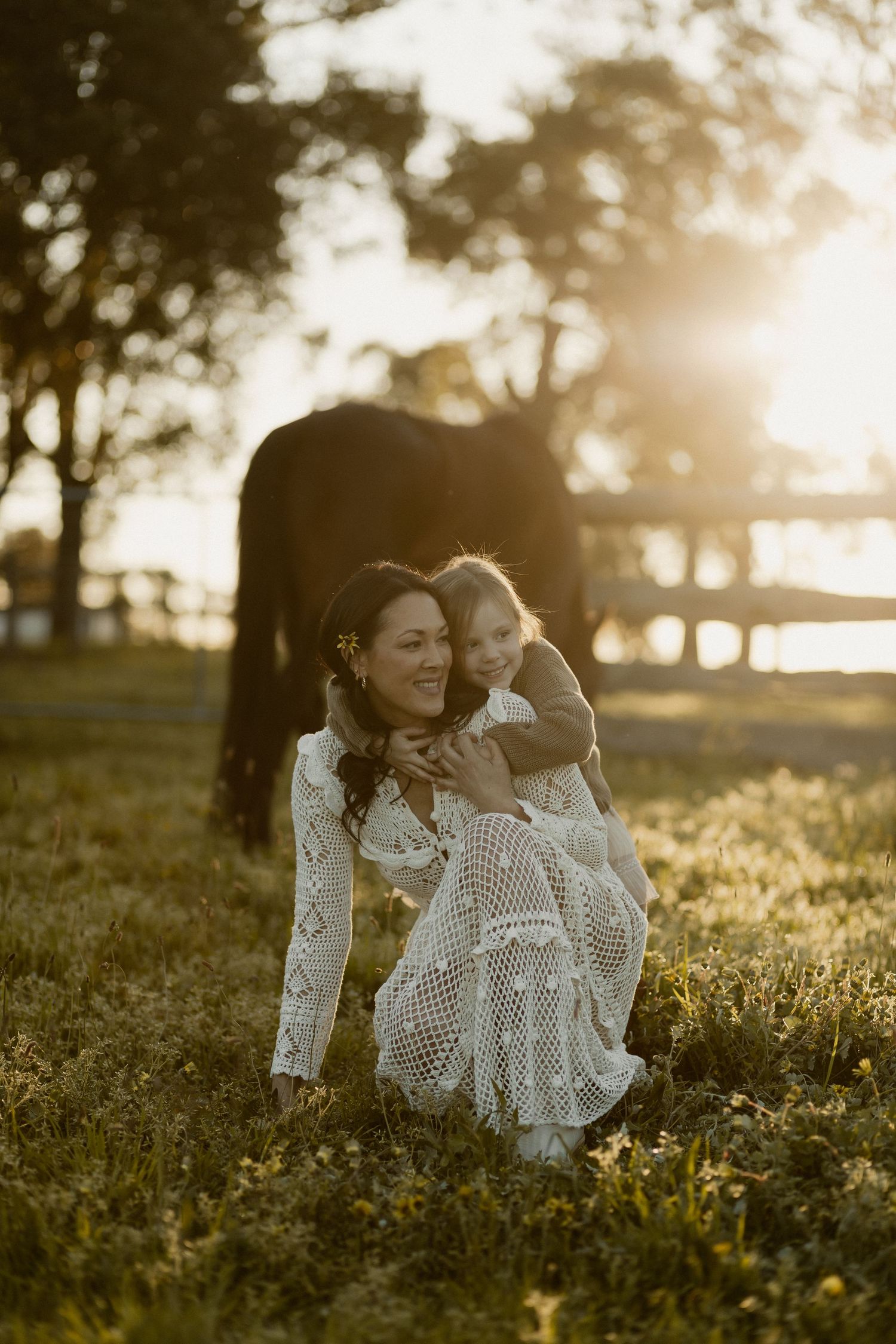A tender moment captured at sunset in a grassy field with sunlight streaming through trees in the background.