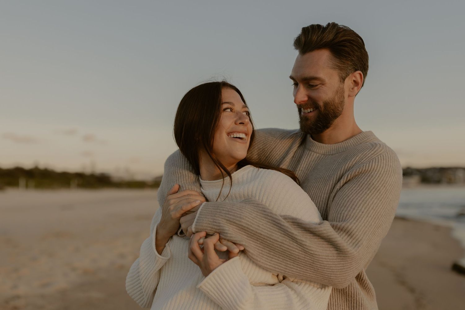A couple in cozy sweaters embraces and laughs together on a beach at sunset.