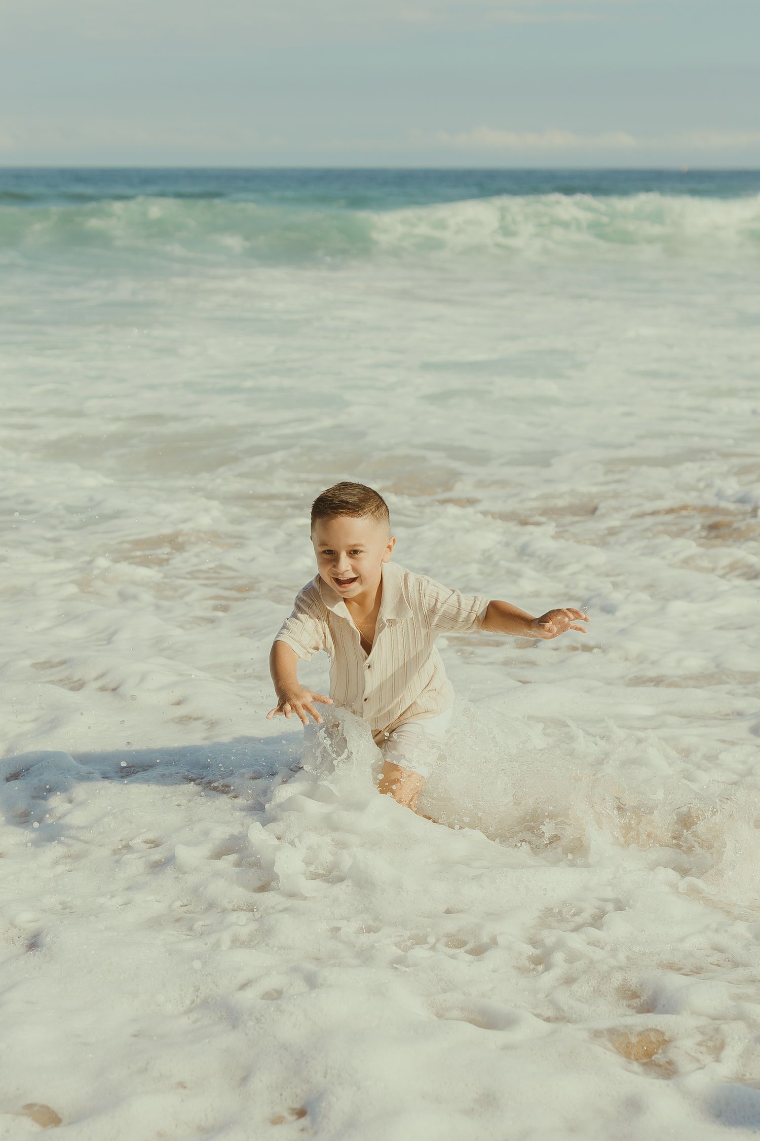 Person in white dress playing and splashing in ocean waves at beach during a seaside photoshoot.