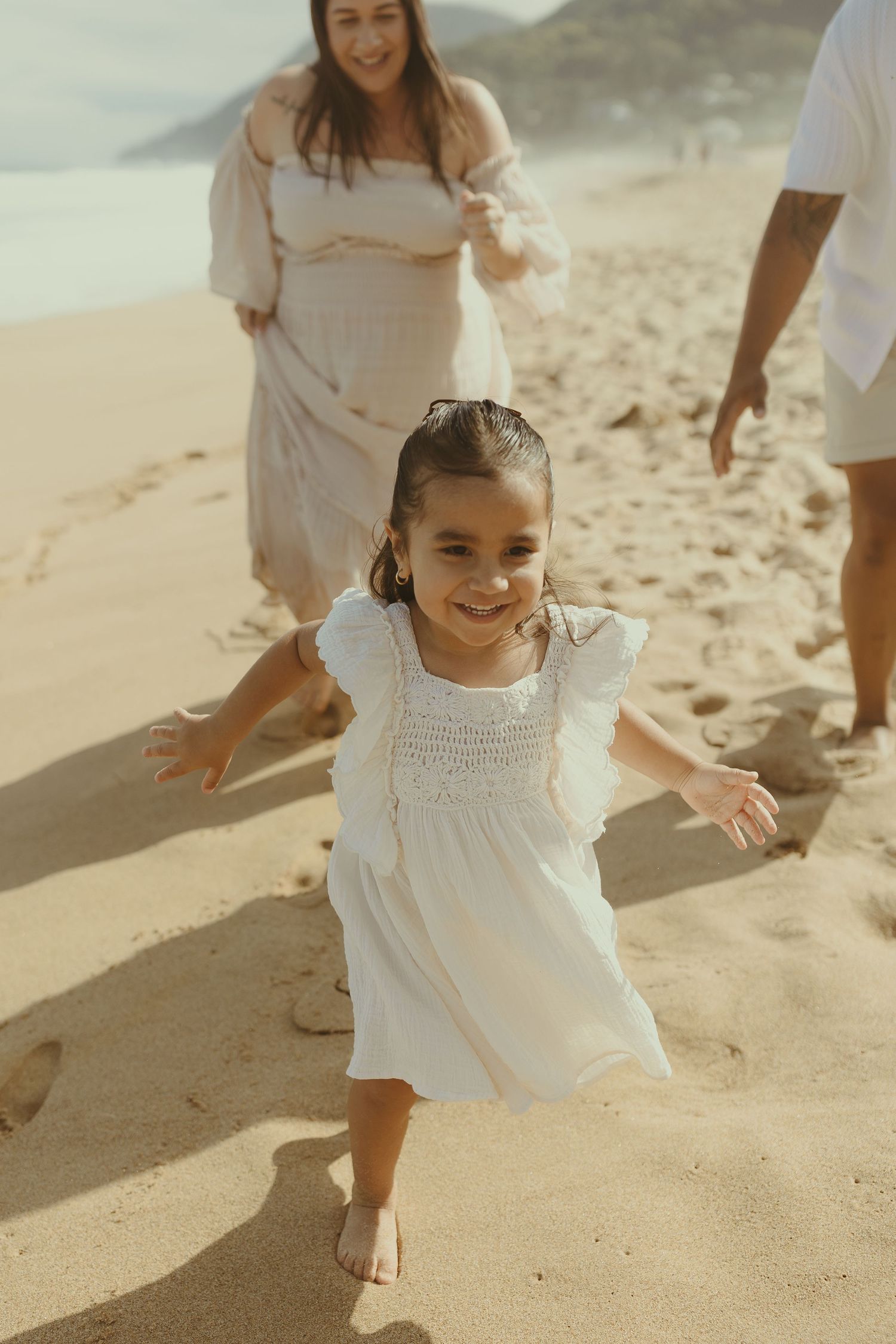 A joyful family wearing white outfits plays together on a sandy beach during golden hour.