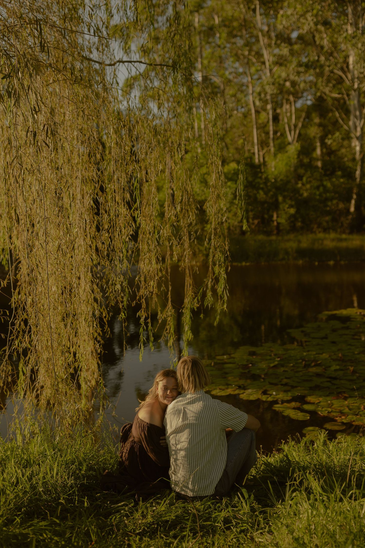 Two people sit close together by a tranquil pond with lily pads under the shade of a weeping willow tree.