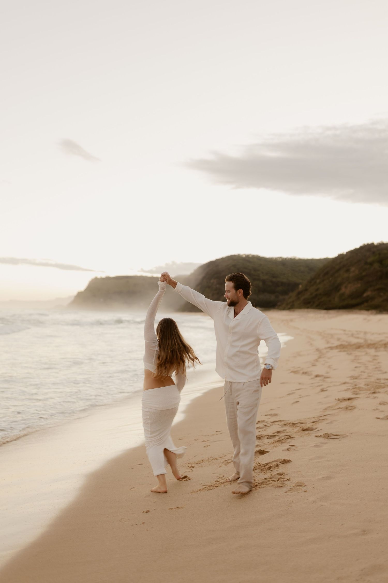 A couple in white clothing dances playfully together on a scenic beach at sunset with mountains in the background.