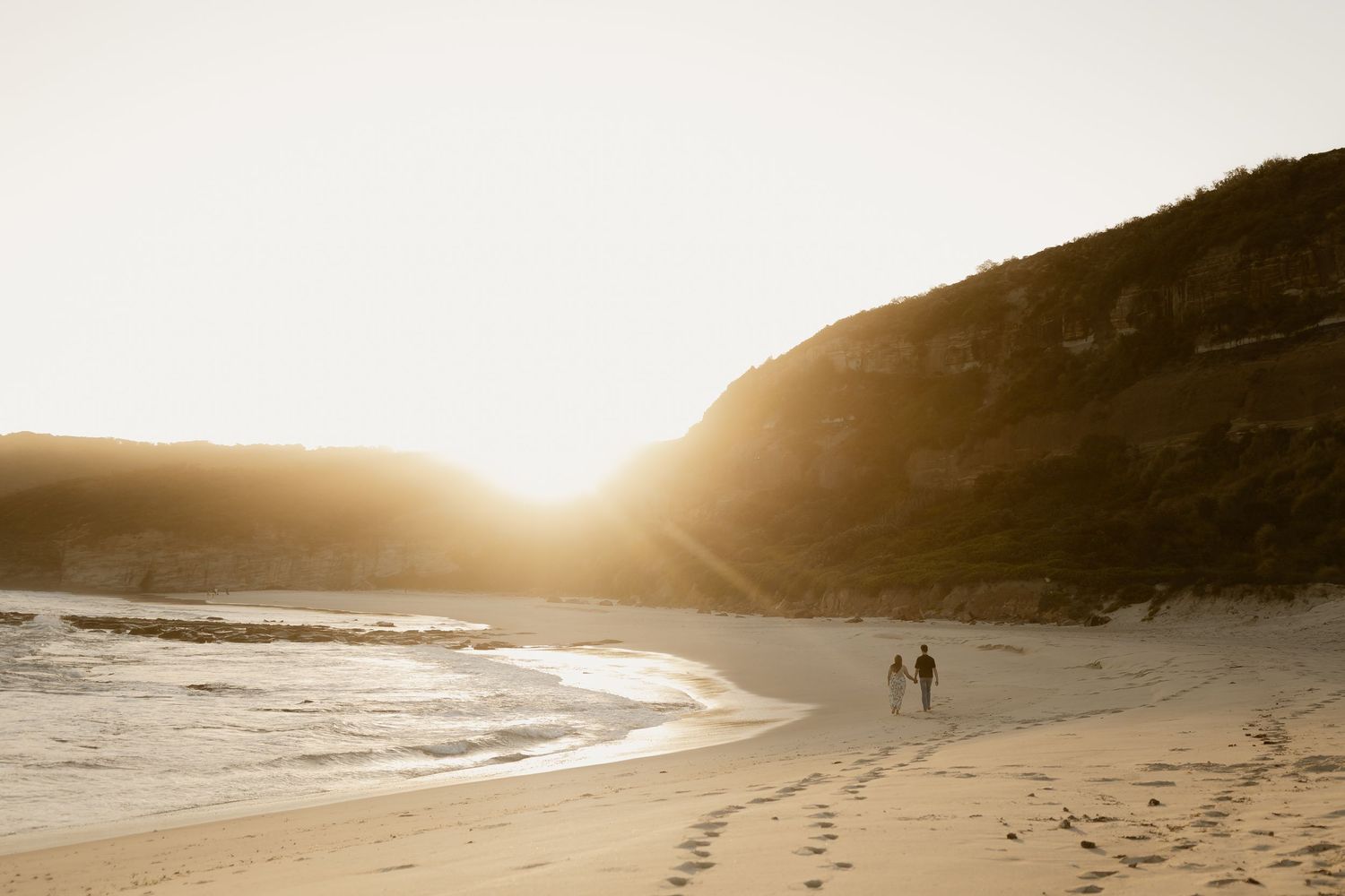 Silhouetted figures walk along a curving beach shoreline at sunset with hills in the distance.