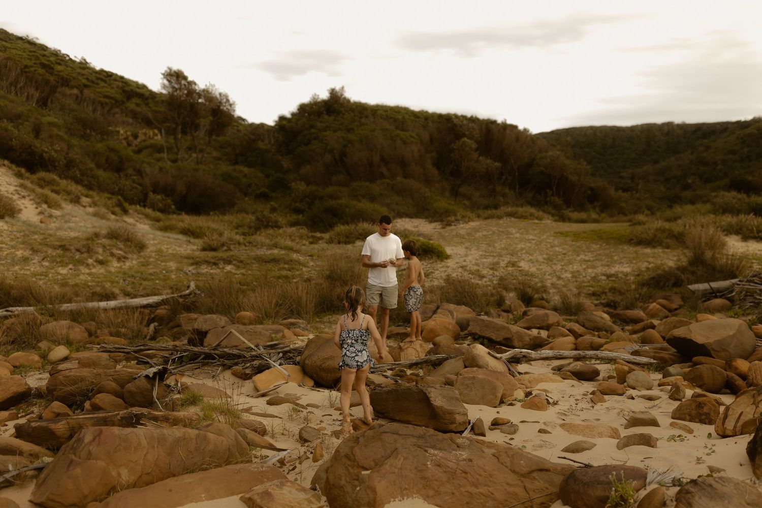 People explore a rocky coastline with mountains and ocean waves in the background.