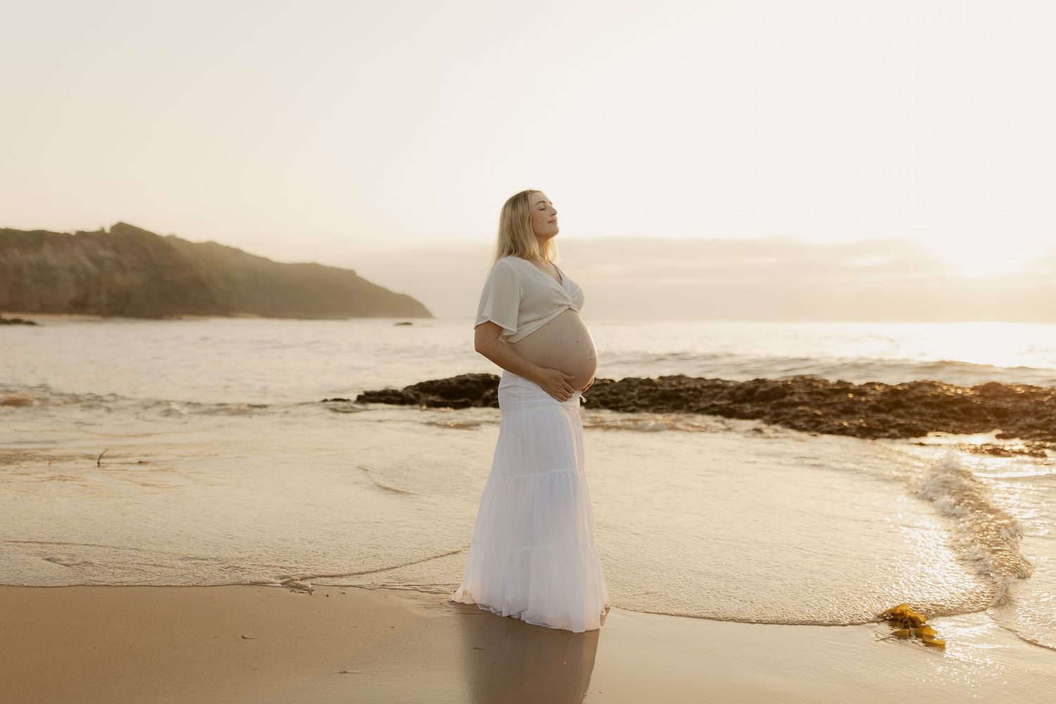 A woman in a white dress walks along a misty beach shoreline at sunset.