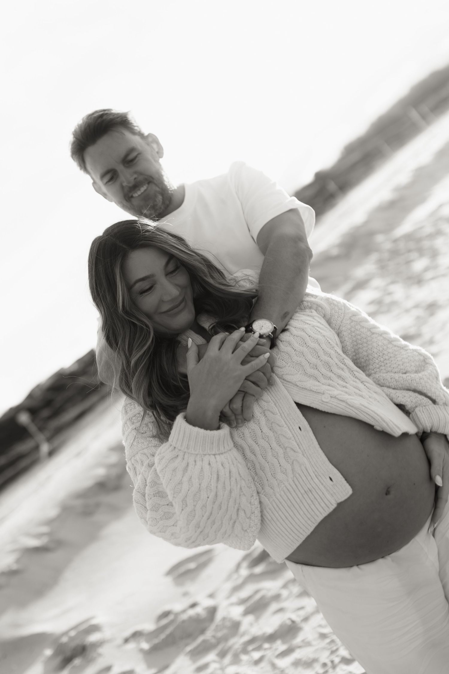 Couple embracing on beach during maternity photoshoot, man hugging pregnant woman from behind, black and white.