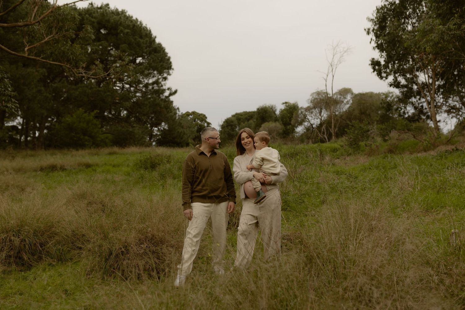 A family of three stands together in a vast green meadow surrounded by trees on an overcast day.