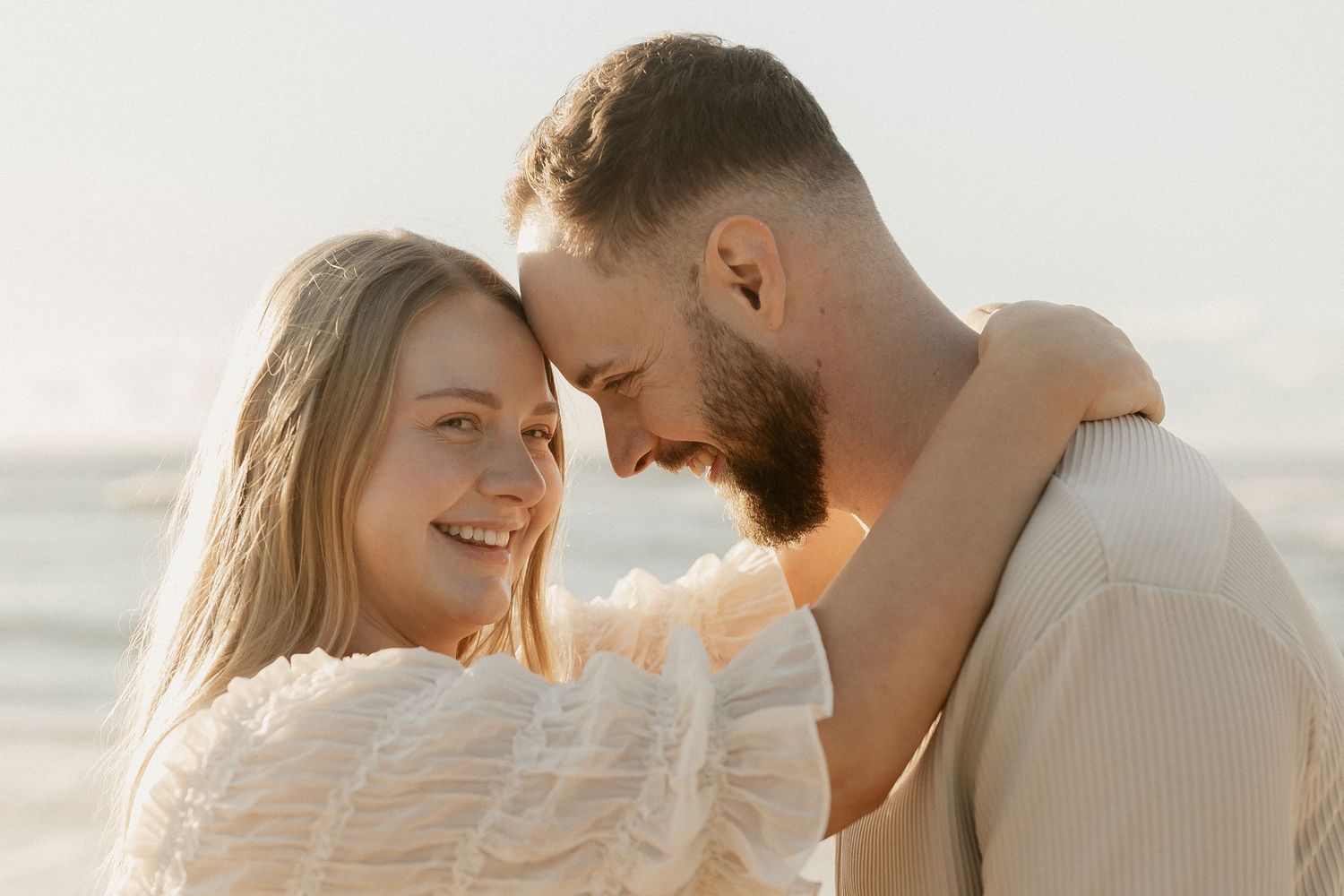 Smiling couple touching foreheads romantically on a beach at golden hour, woman in white wrap.