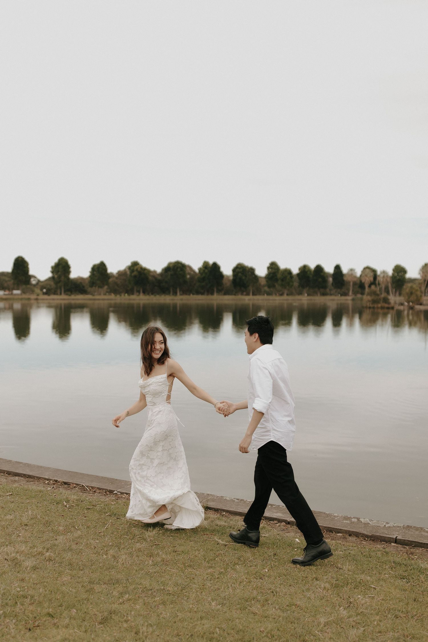 A couple playfully runs along the edge of a serene lake surrounded by trees at sunset.