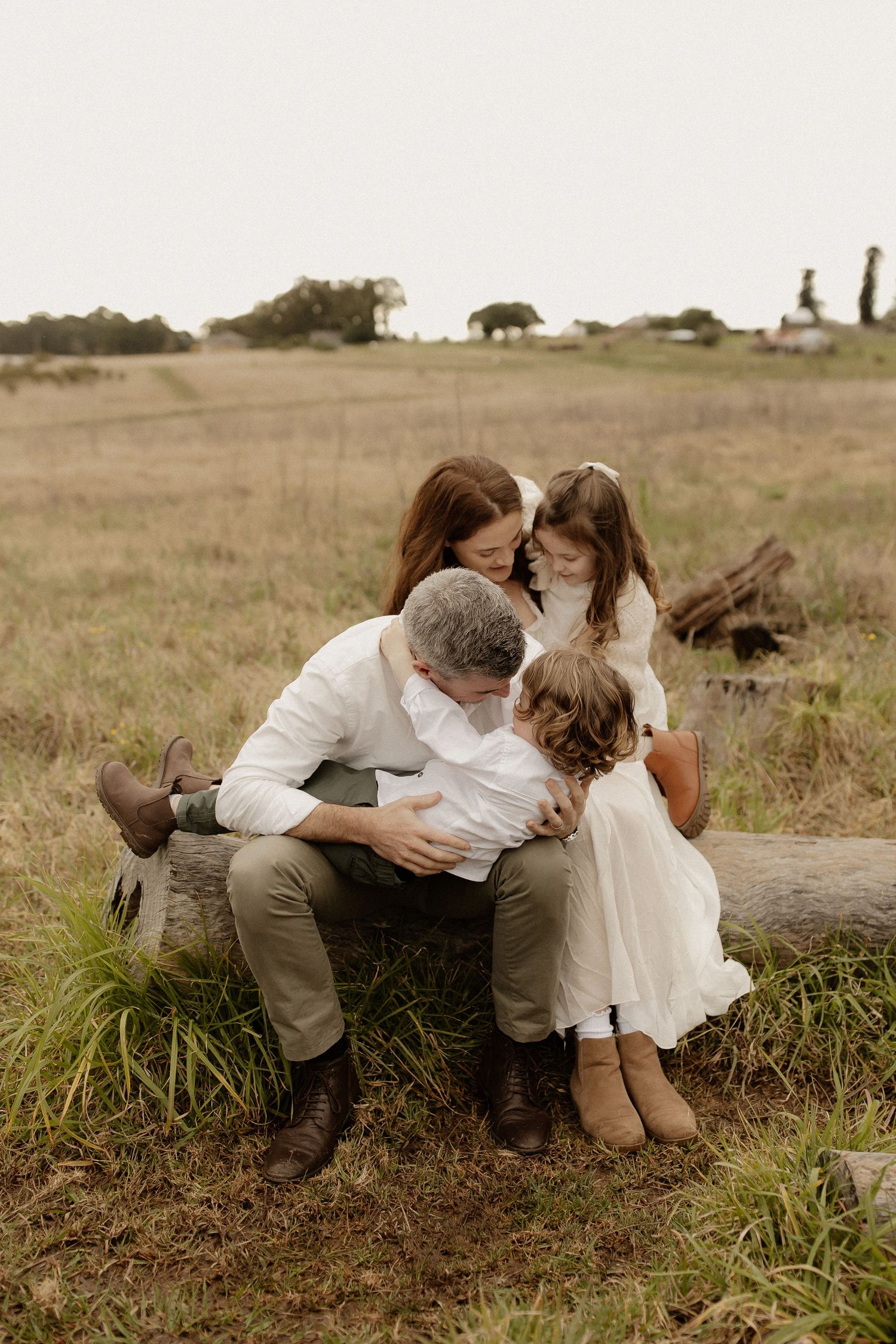 A romantic couple in white clothing shares tender moments while sitting in a grassy field at sunset.
