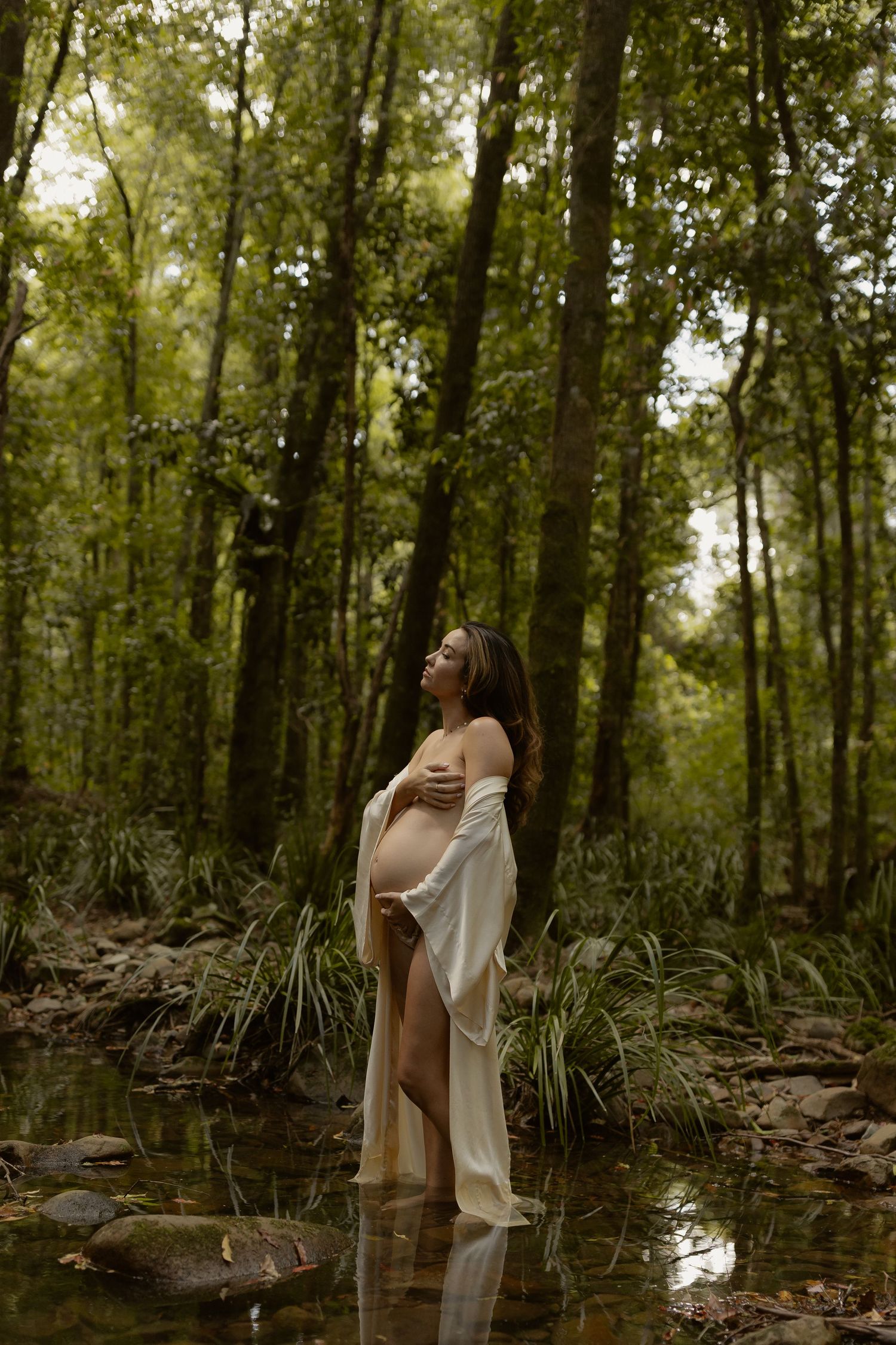 A pregnant woman in a white wrap stands peacefully in a tranquil forest pond surrounded by tall trees and lush foliage.
