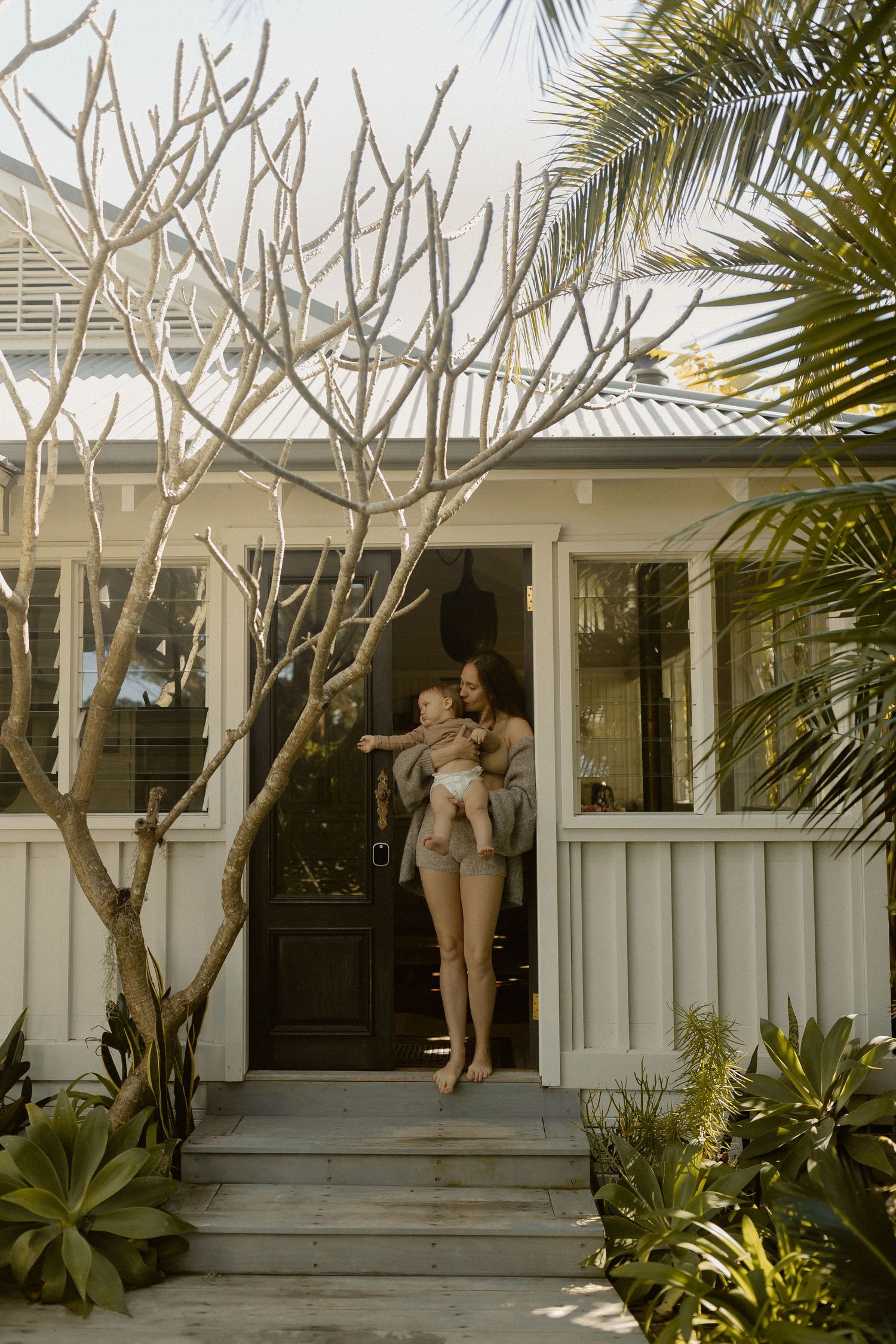 Ethereal figures move across a sun-drenched porch with palm trees and white architectural details creating a dreamy scene.