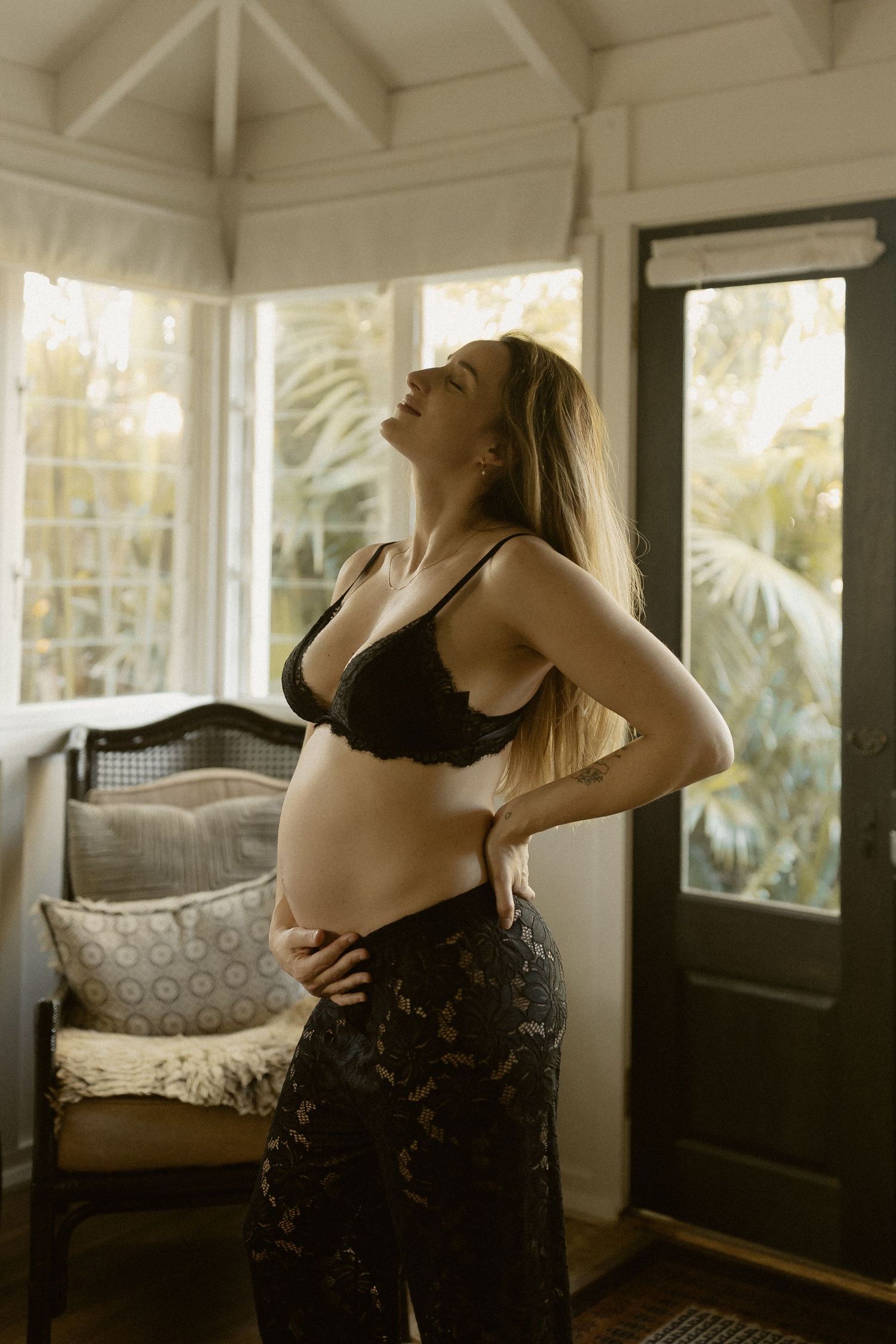Maternity photo of expectant mother in black lingerie standing in sunlit room with tropical plants visible through windows.
