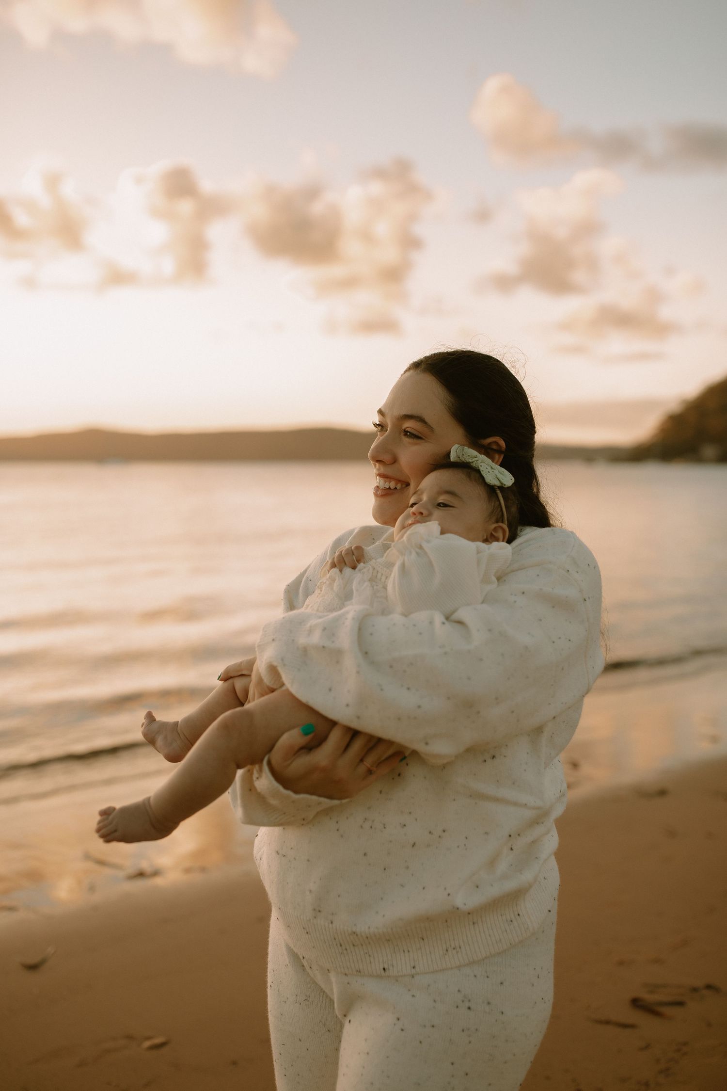 A person in a cozy white sweater dances freely on a golden sunset beach with waves and mountains in the background.