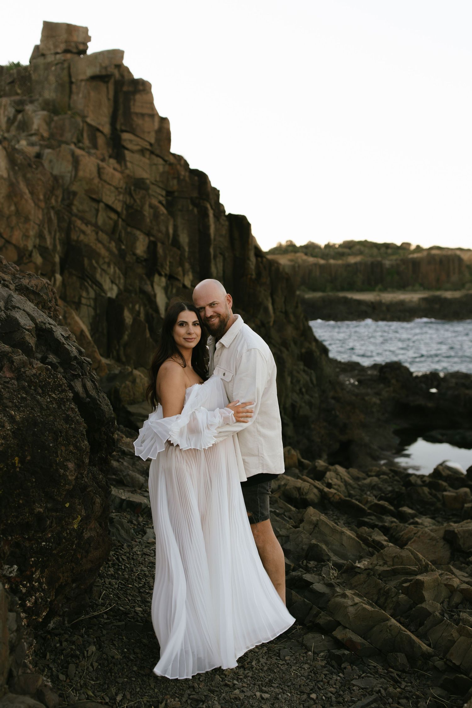 Couple embracing on dramatic rocky shoreline in flowing white attire.