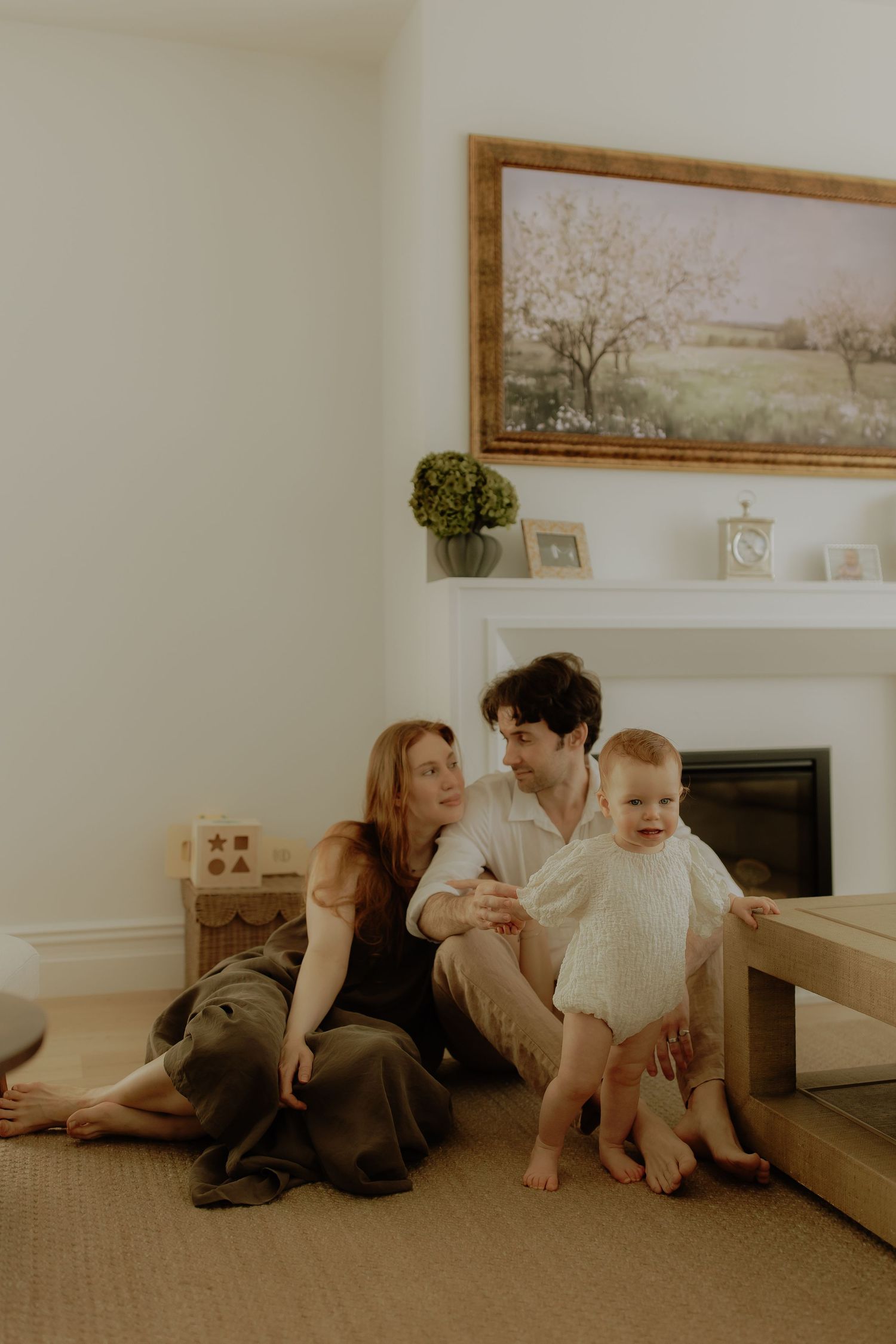 A young family sits on the floor by a fireplace, parents smiling at their happy baby in a cozy living room.