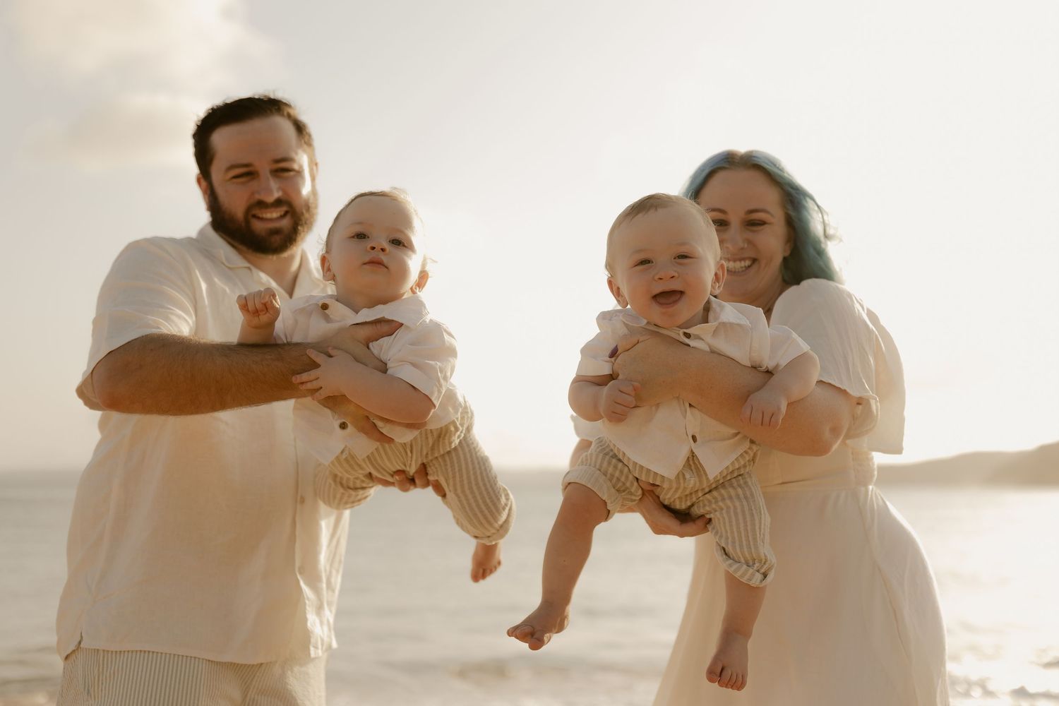 A couple wearing white outfits holds twin babies while standing on a beach during golden hour.