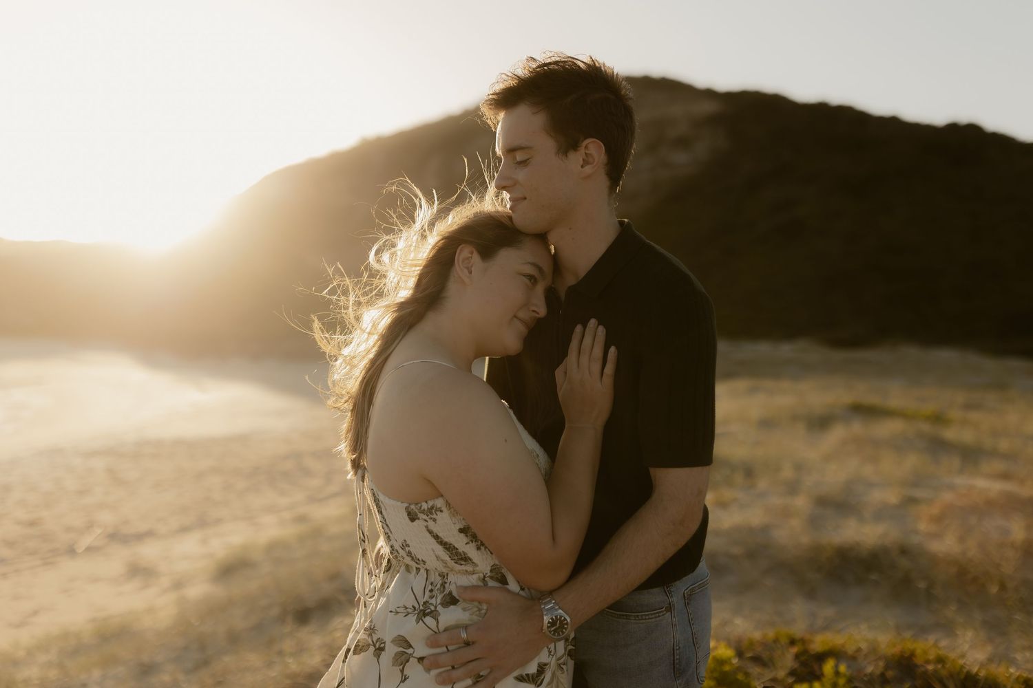 A couple embraces during golden hour on a hillside with sunlight creating a warm backlit glow in the scene.
