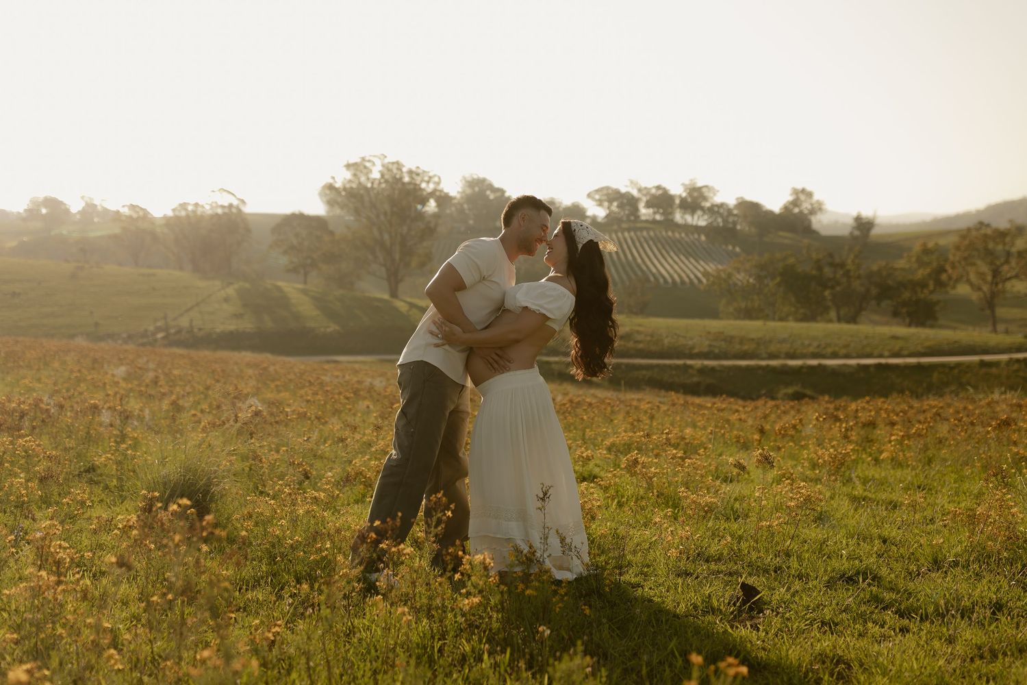 A romantic couple embraces in a sunlit meadow with golden light streaming through trees in the background.