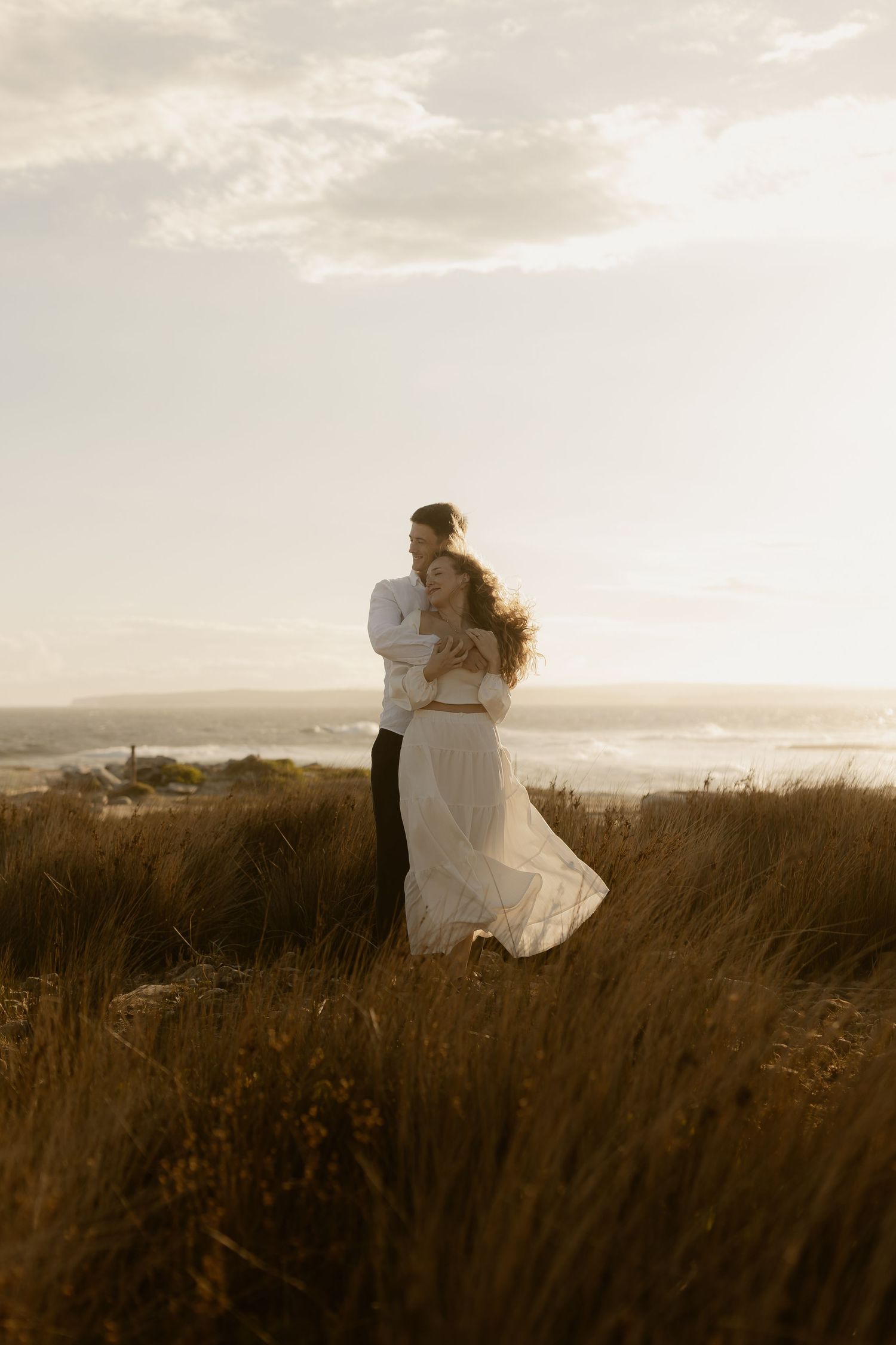 Two figures embrace in a grassy field overlooking the ocean at sunset.