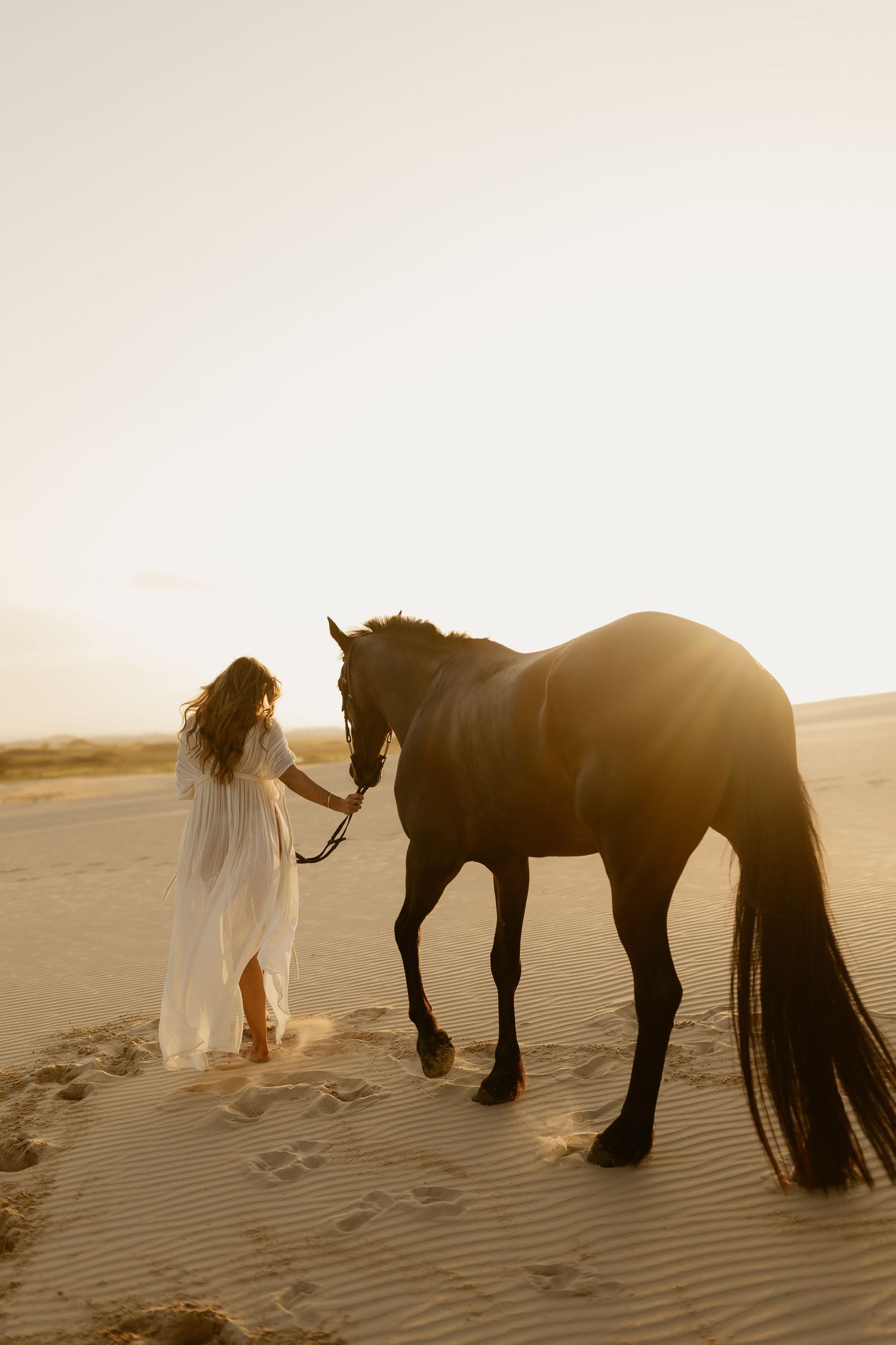 Woman in white dress leads dark horse across sandy dunes bathed in warm golden sunset light.