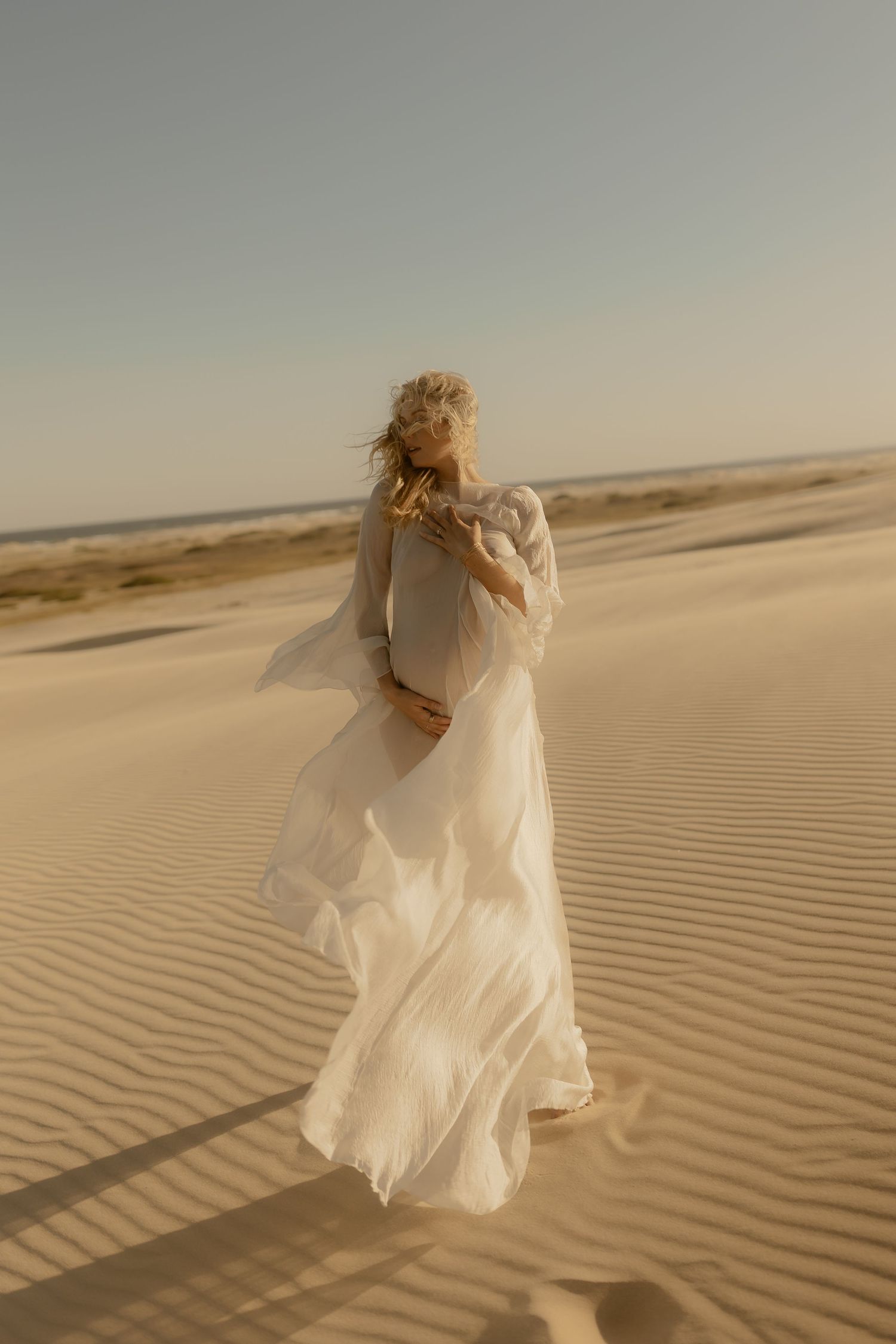 Blonde woman in flowing white dress standing on sandy desert dunes with wind blowing her gown.