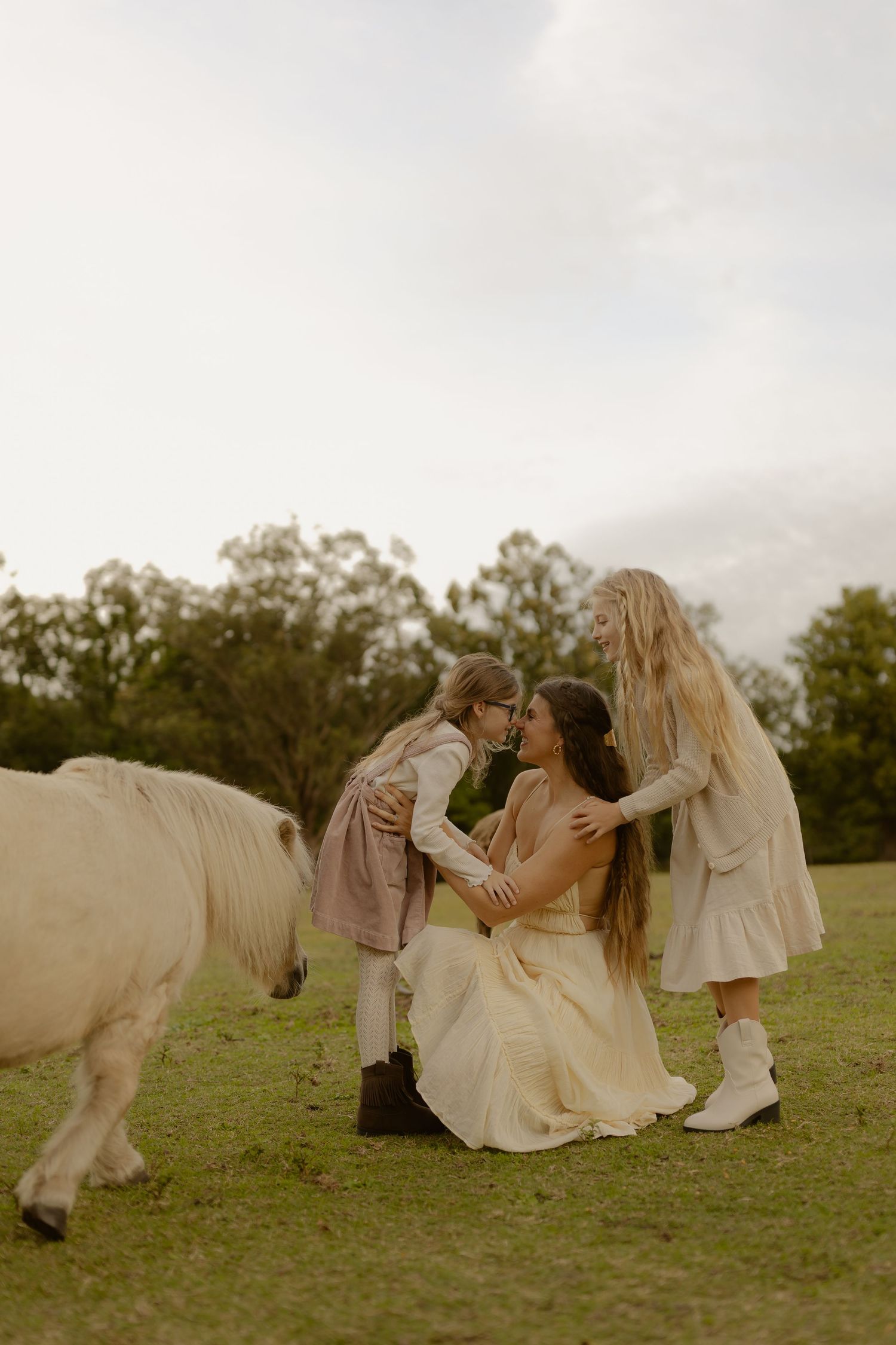 A mother kneels on grass sharing a sweet moment with two daughters near a white pony.