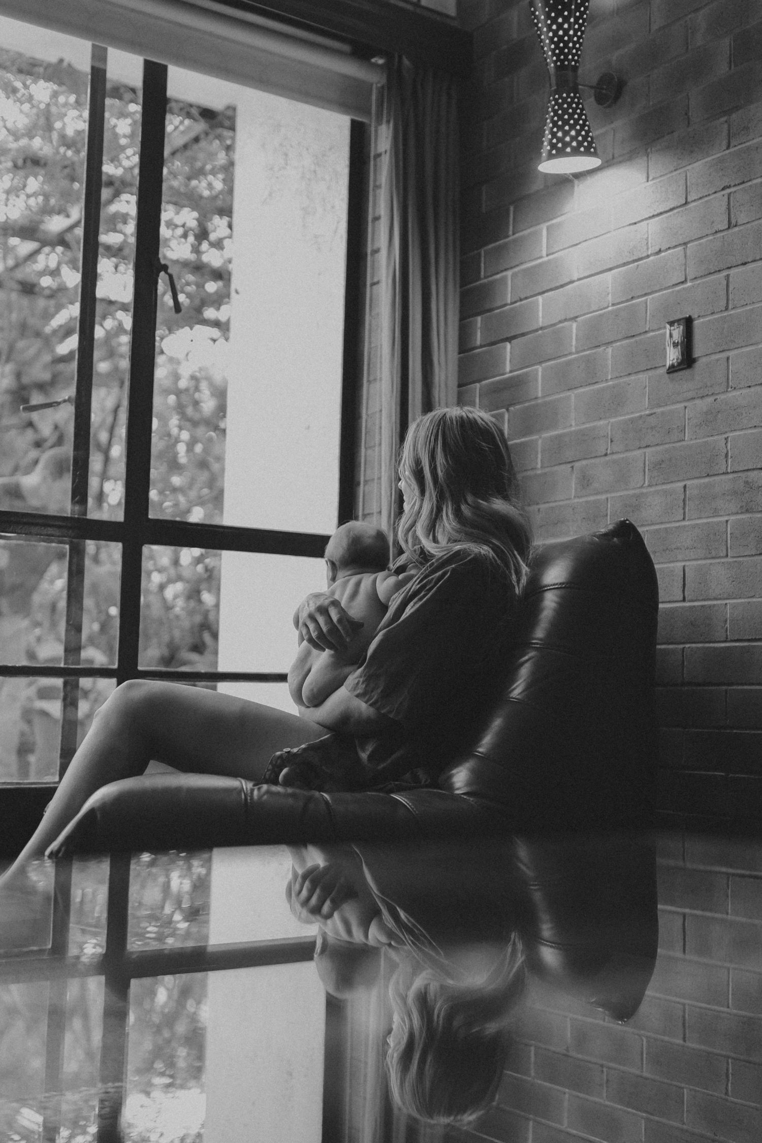 Black and white photo of mother cradling newborn in leather chair by large window.