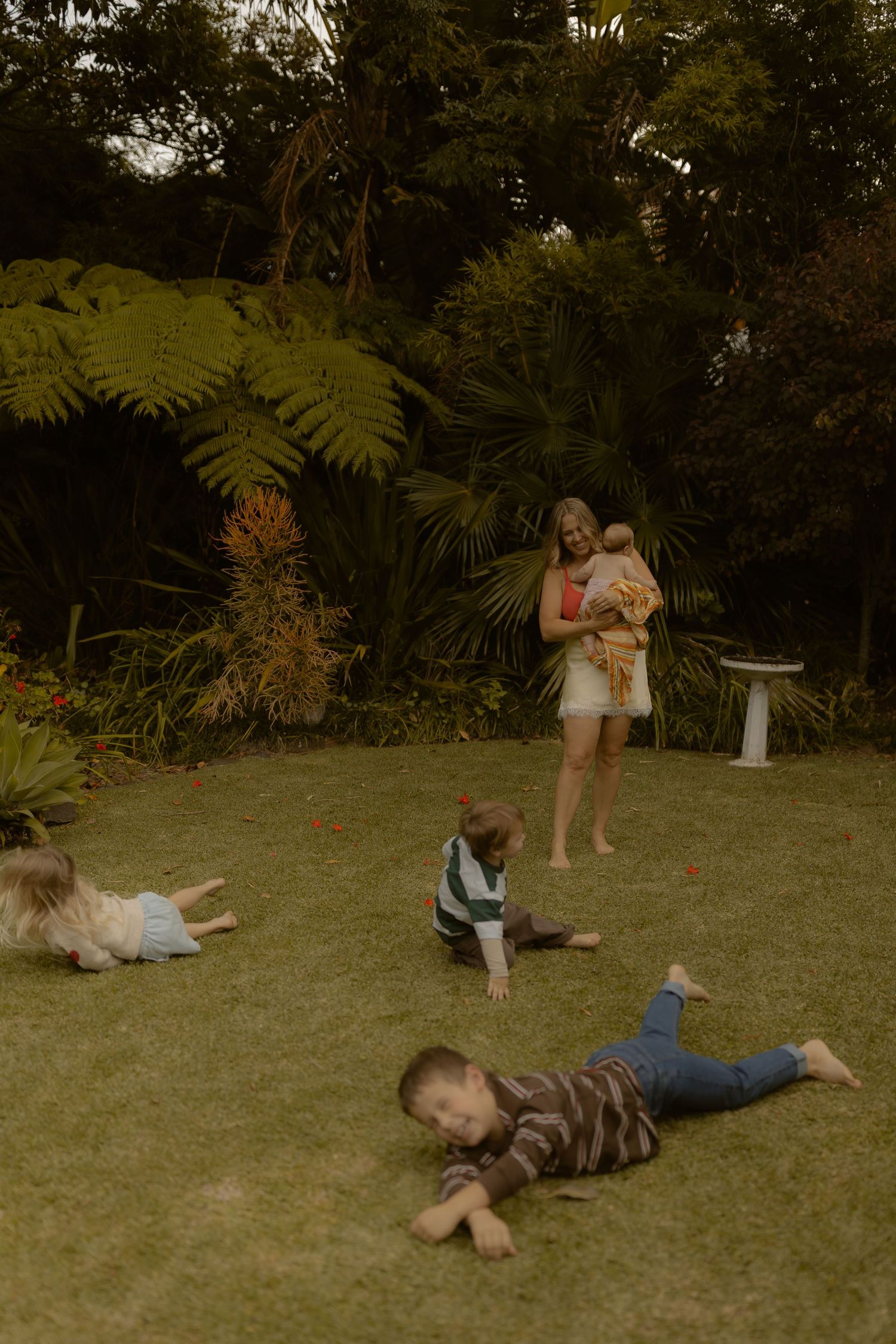 Mother holding baby watching three young children playing on green lawn in lush garden.