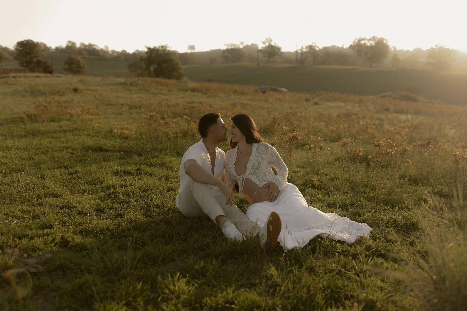 Couple in white outfits sitting on green grass field at golden hour sunset, sharing a romantic moment.