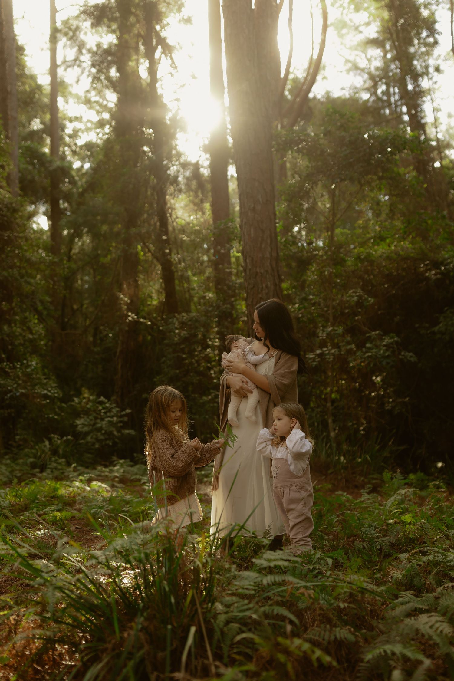 Mother holds newborn while two young children stand among lush forest ferns in golden sunlight.