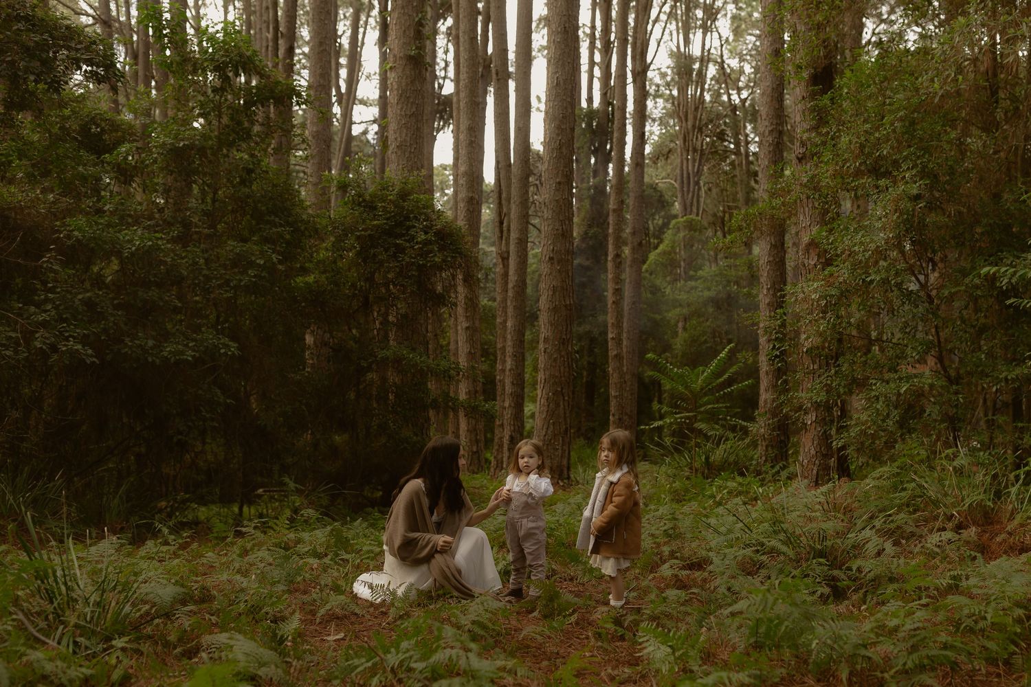 A mother and two children sit together in a lush green forest surrounded by tall trees and ferns.