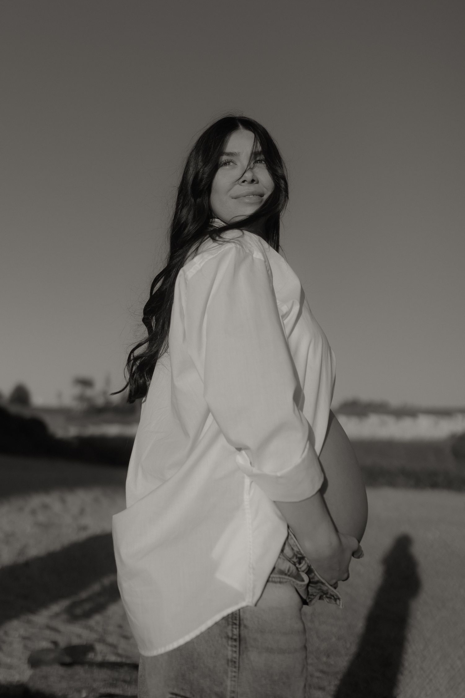 Black and white portrait of pregnant woman in white shirt looking over shoulder outdoors.