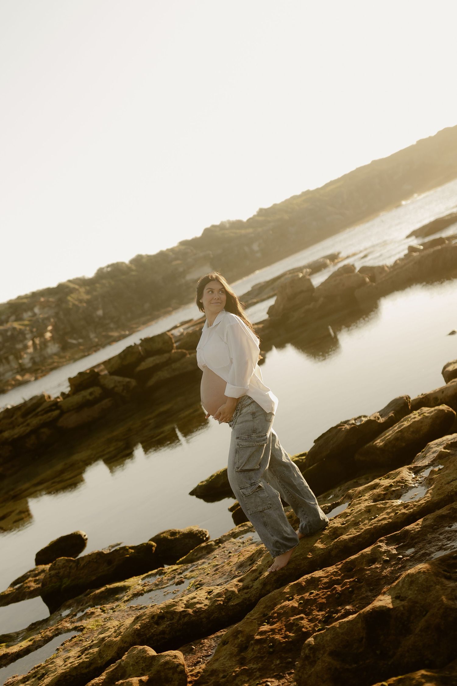 Pregnant woman in white shirt and jeans standing on rocky shoreline at golden hour sunset.