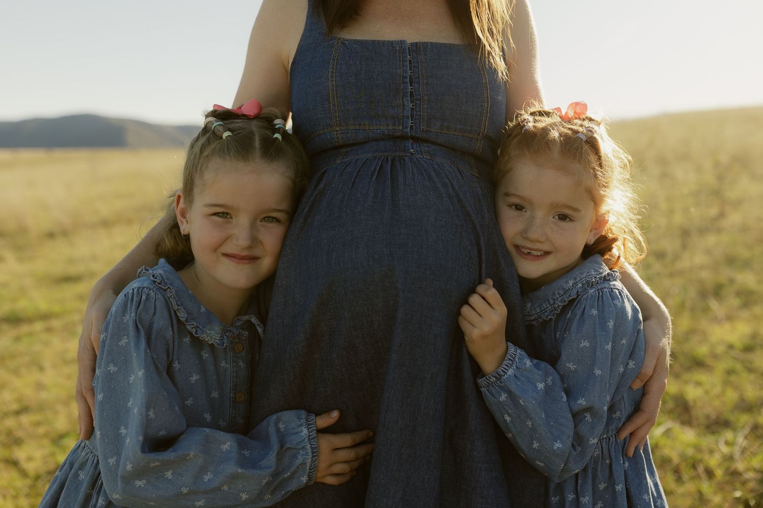Two young girls in denim dresses hugging their pregnant mother outdoors in a sunny field.