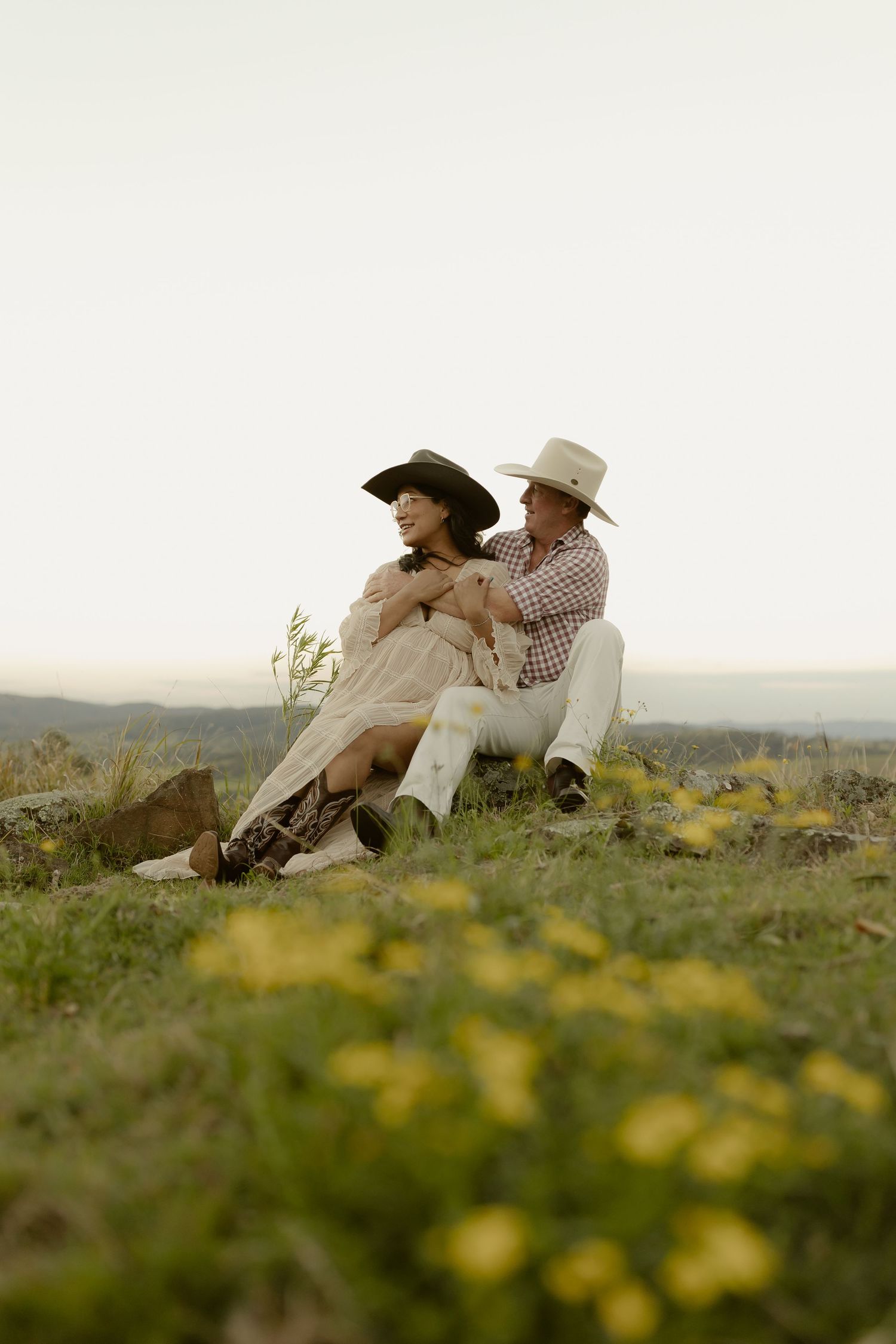 A couple in cowboy hats sit together on a grassy hill at dusk, embracing romantically with a scenic landscape behind them.