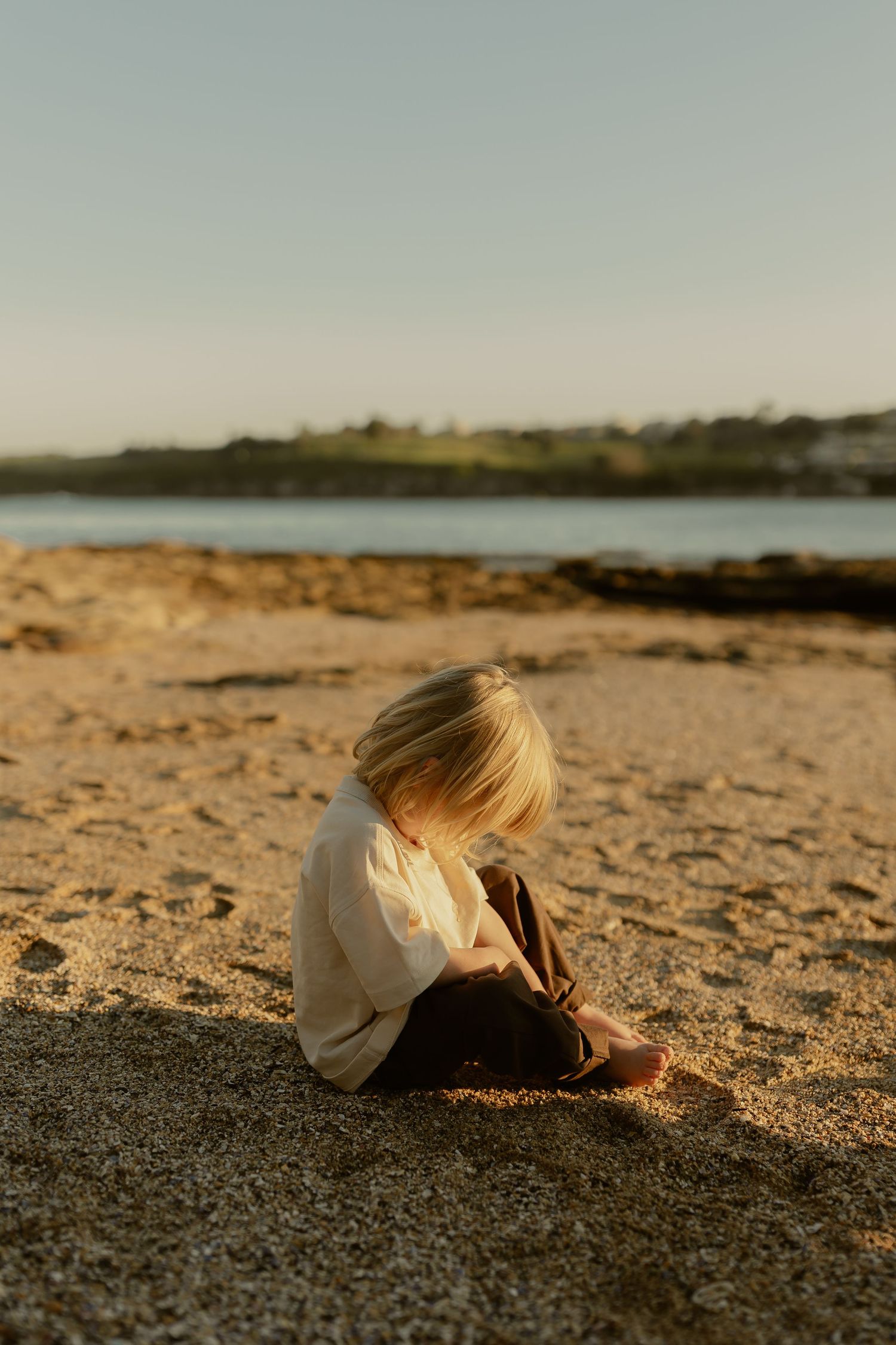 A blonde toddler sits alone on sandy beach at golden hour, looking down at the sand near the water.