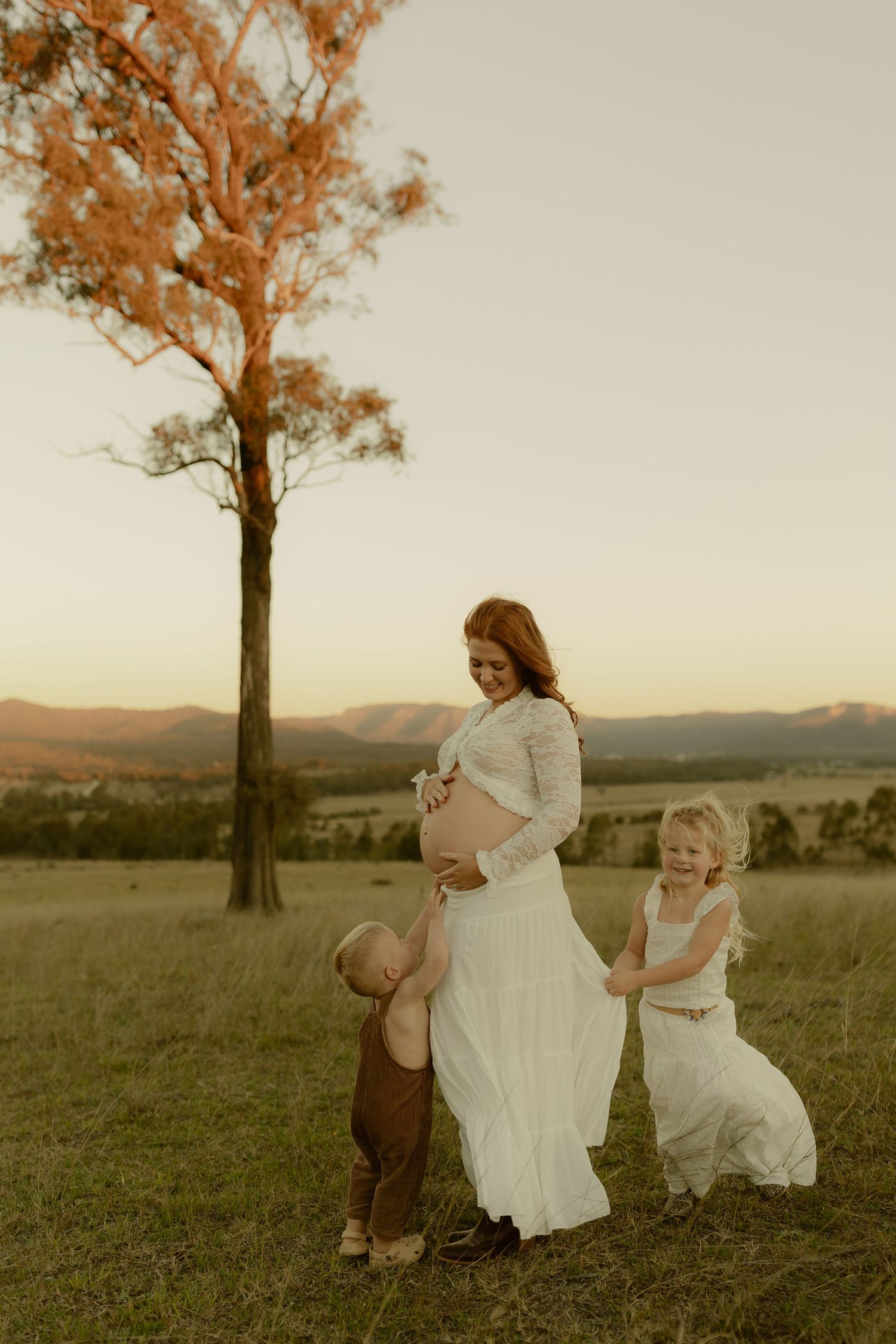 Pregnant woman in white lace dress with two young children in golden field at sunset.