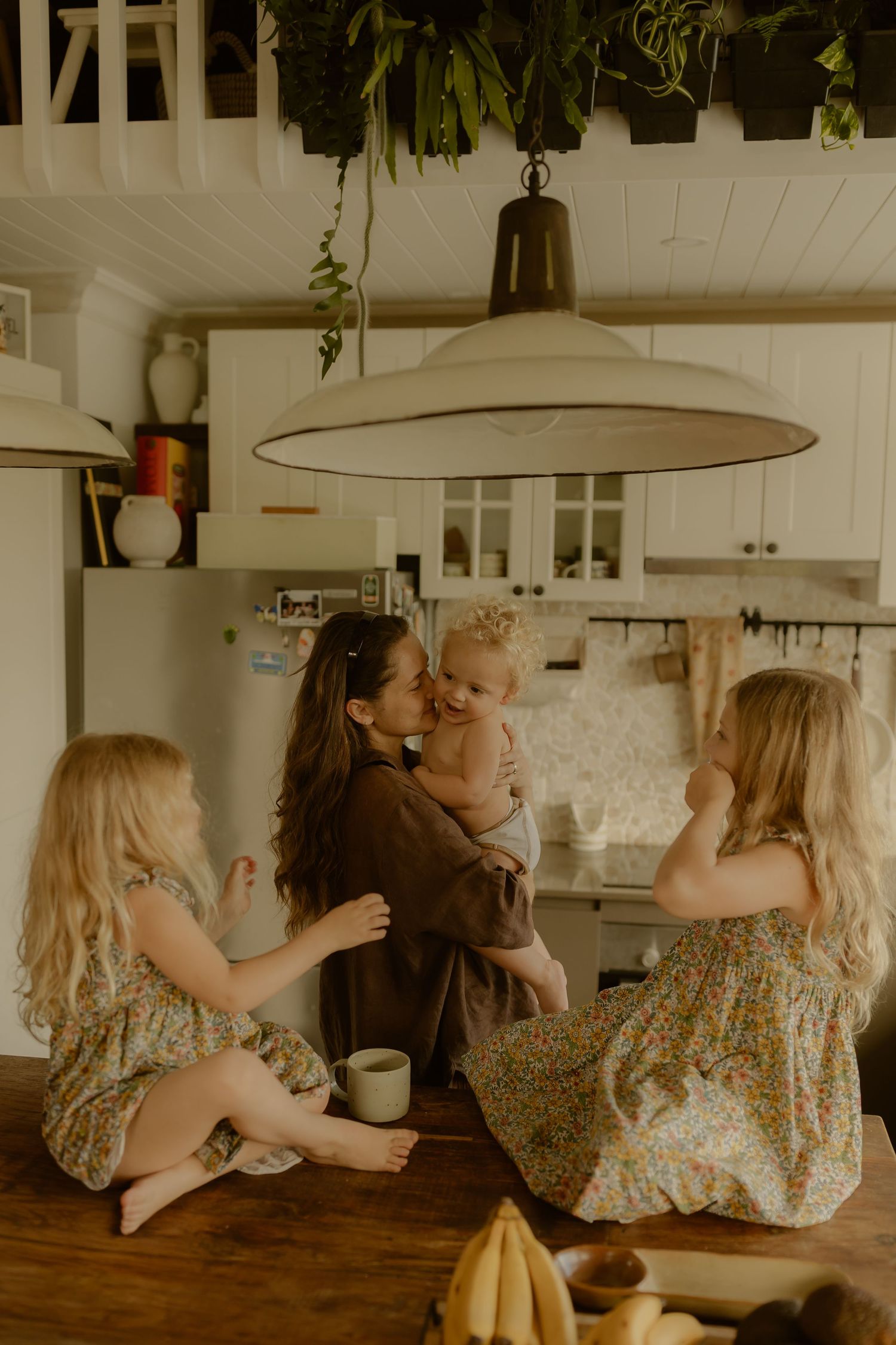 Mother holding baby while two blonde girls sit on kitchen counter in bright farmhouse kitchen.