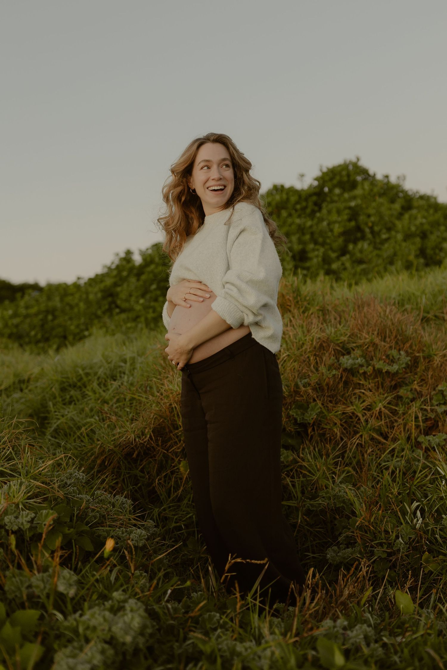 Smiling pregnant woman in white sweater and black pants cradling her baby bump in a lush green field at golden hour.