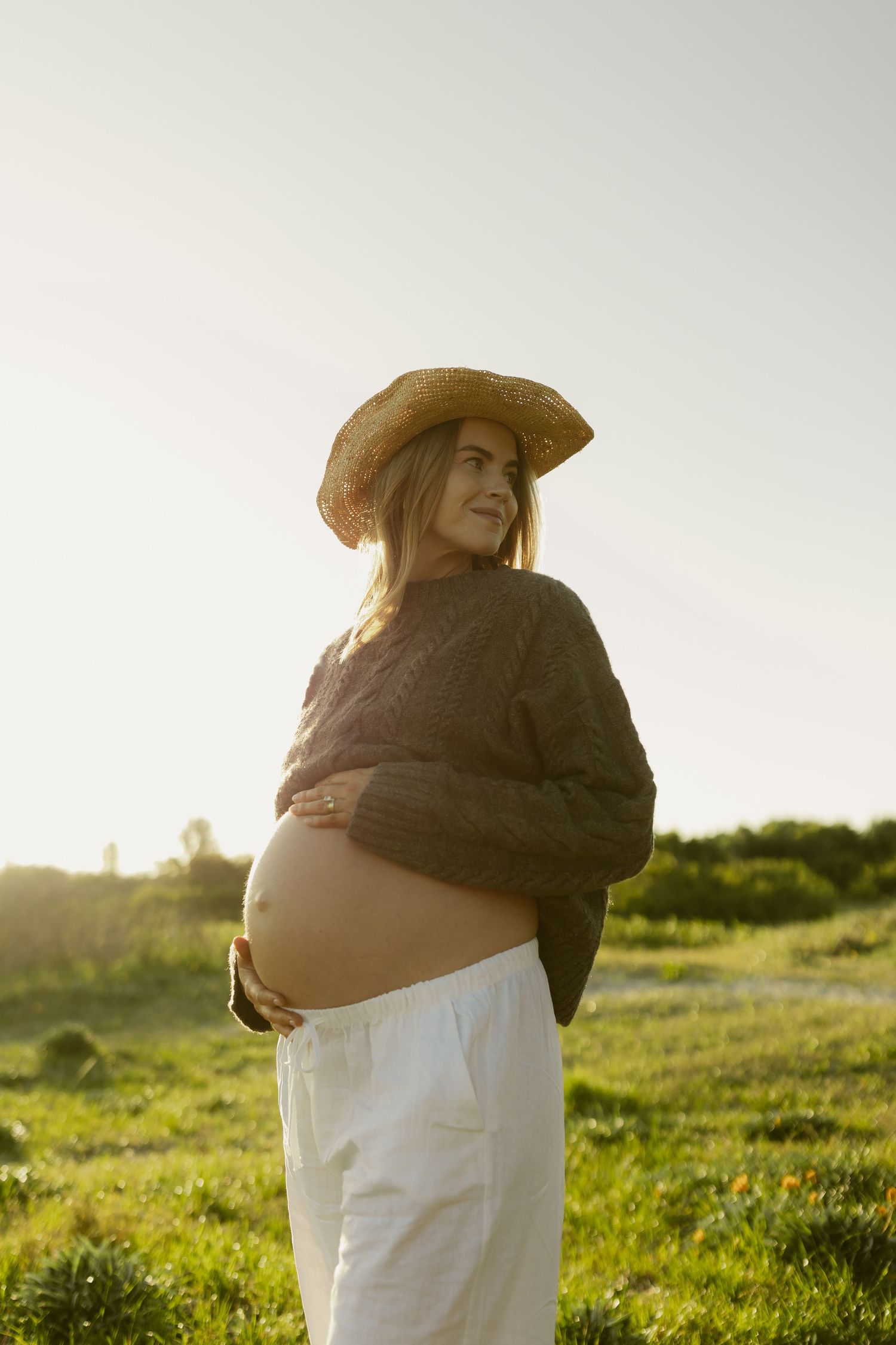 Pregnant woman in straw hat and brown sweater holding baby bump in golden sunset field.