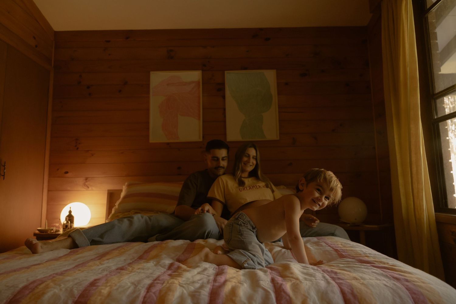 A couple and child relax together on a bed in a cozy warm-toned wood-paneled bedroom.