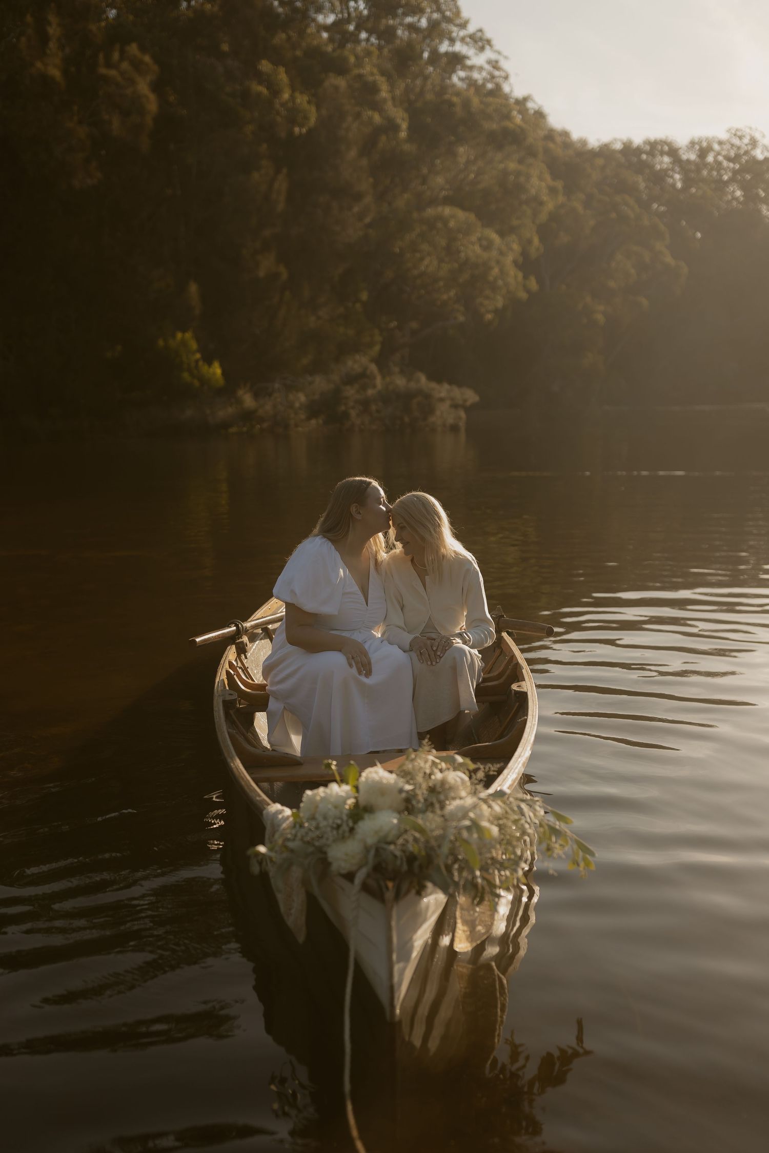 Two brides in white dresses share a kiss in a flower-adorned wooden canoe on a serene lake at golden hour.